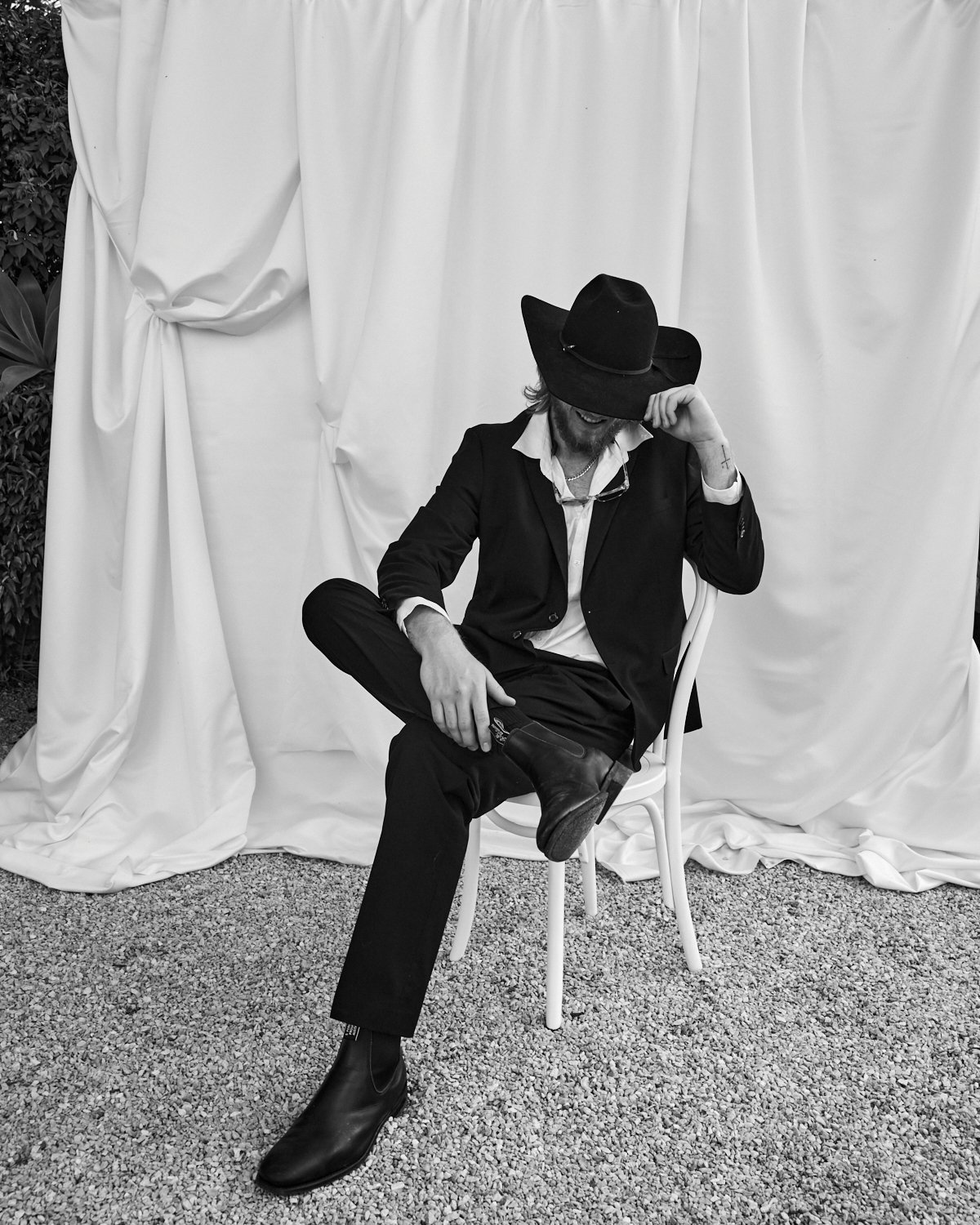 A man dressed in a black suit with a white shirt, wearing a large black hat, sitting on a chair on a gravel surface with a white draped fabric backdrop. He is smiling, with his right arm resting on his raised right knee, and his left hand holding the