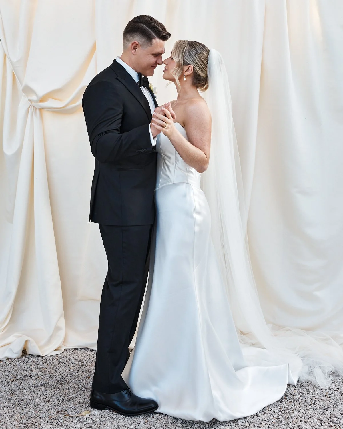 A bride and groom share a tender dance, gazing lovingly at each other indoors with cream-colored curtains in the background. The bride wears a white strapless wedding gown and veil, while the groom wears a black tuxedo.