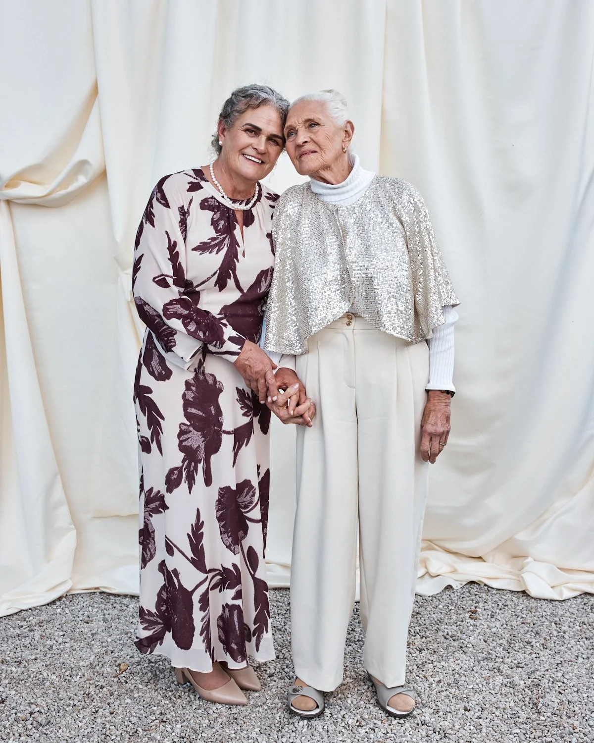 Two elderly women standing held hands, smiling, in front of a light-colored fabric backdrop.