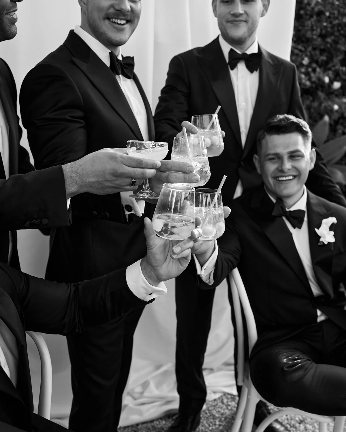 A group of men in tuxedos at a celebration, holding up drinks and smiling, with some seated and some standing.