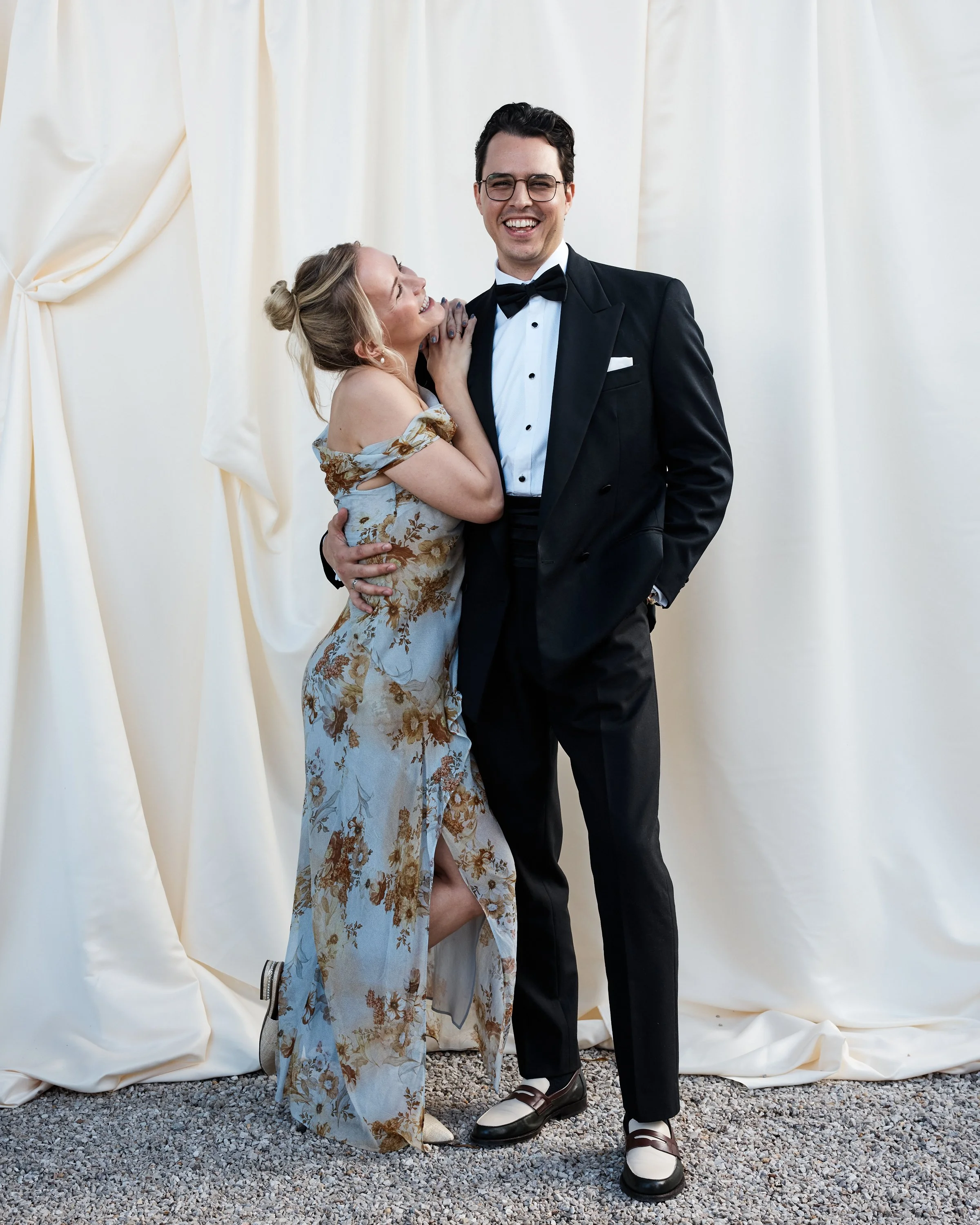A man in a tuxedo and a woman in a floral dress pose together, smiling, against a cream-colored backdrop.