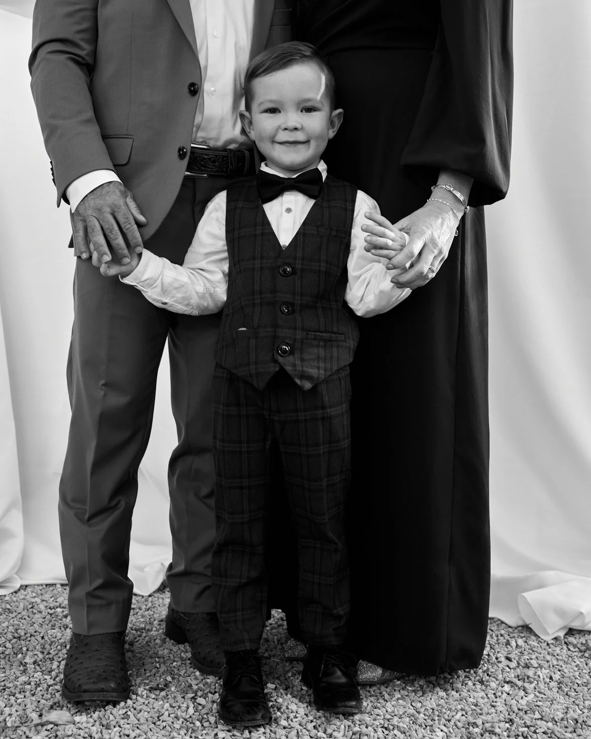 A young boy dressed in a white shirt, bow tie, and plaid vest and pants standing between two adults, who are holding his hands. The adults are dressed in suits. The image is in black and white.