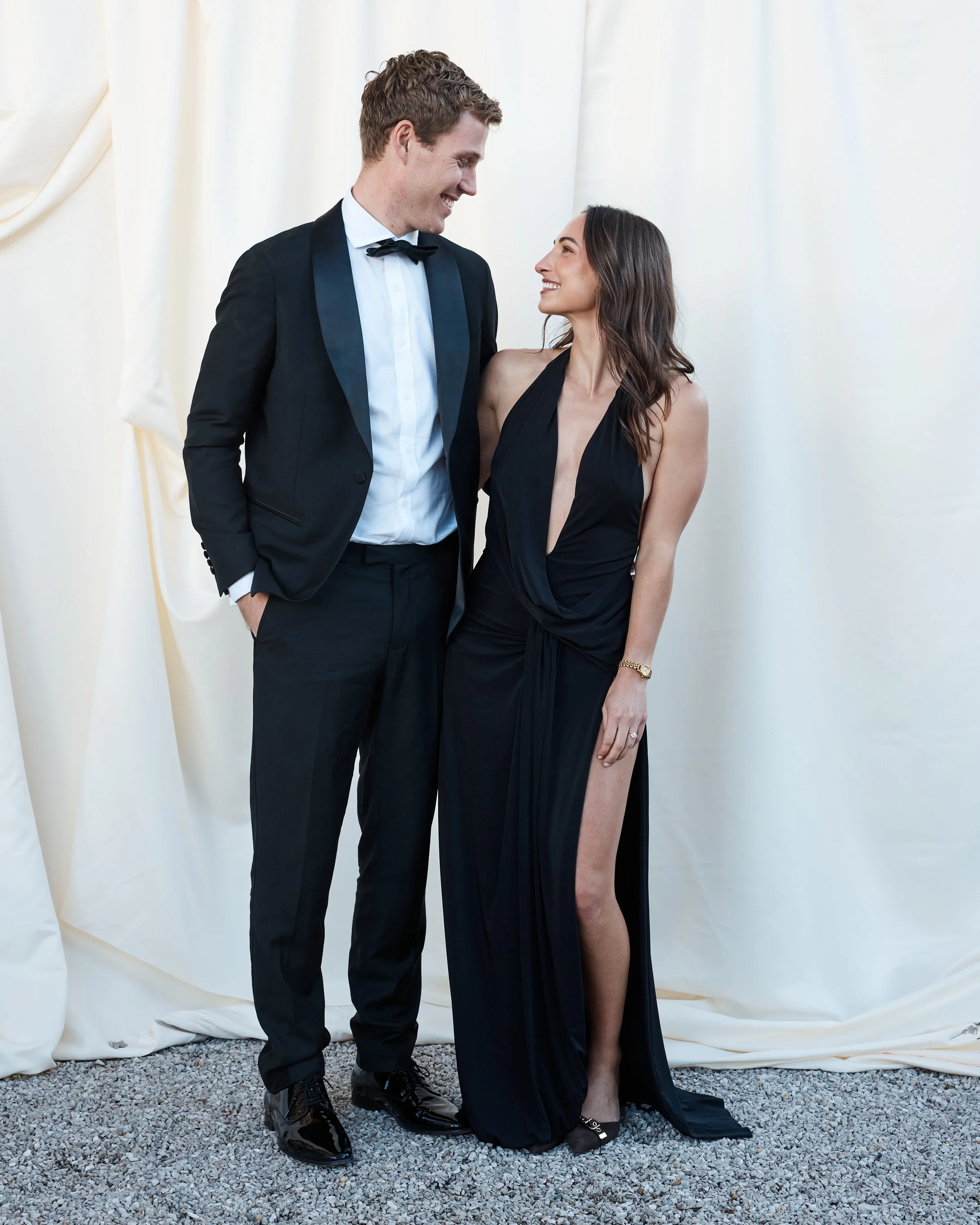 A man in a tuxedo and a woman in a black gown are standing close together, smiling and looking into each other's eyes, against a cream-colored curtain backdrop.