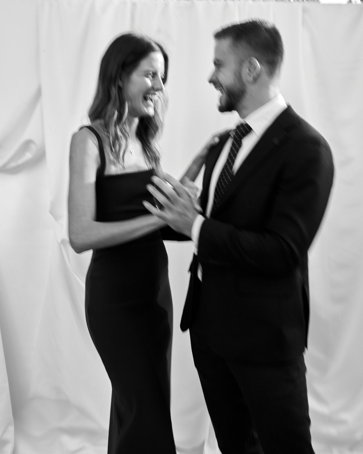 A woman in a sleeveless dress is smiling and touching a man's chest, who is dressed in a suit, as they look at each other happily against a plain background.