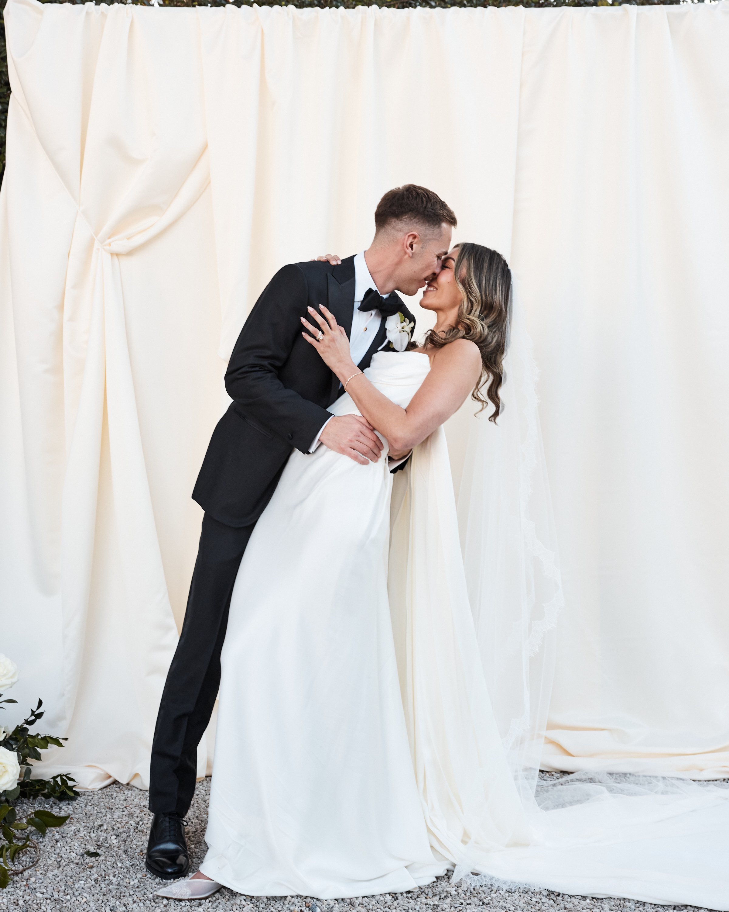 A bride and groom share a kiss during their wedding ceremony, with a cream-colored curtain backdrop.