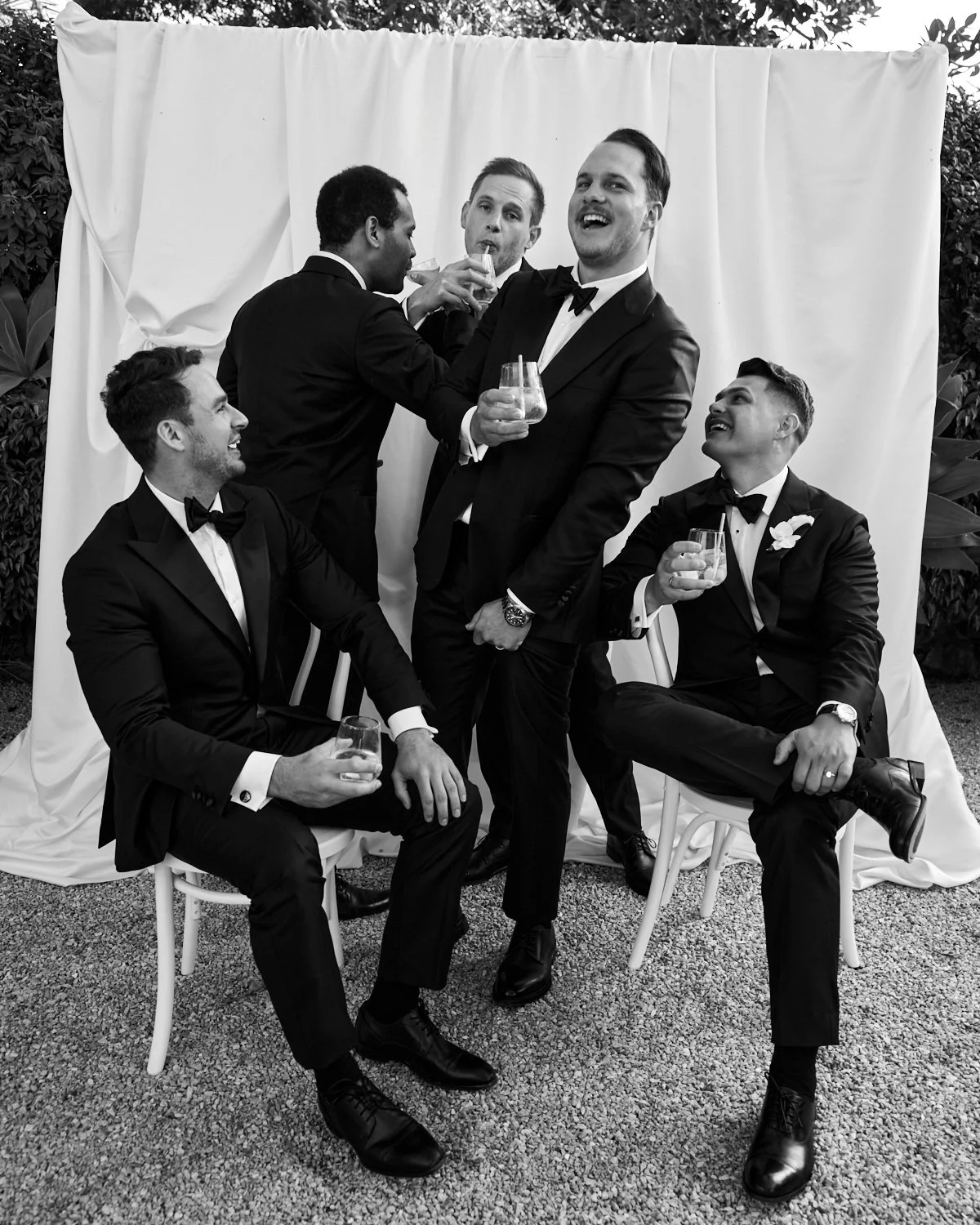 Six men in tuxedos celebrating and enjoying drinks at a formal event, with a white backdrop and outdoor greenery.