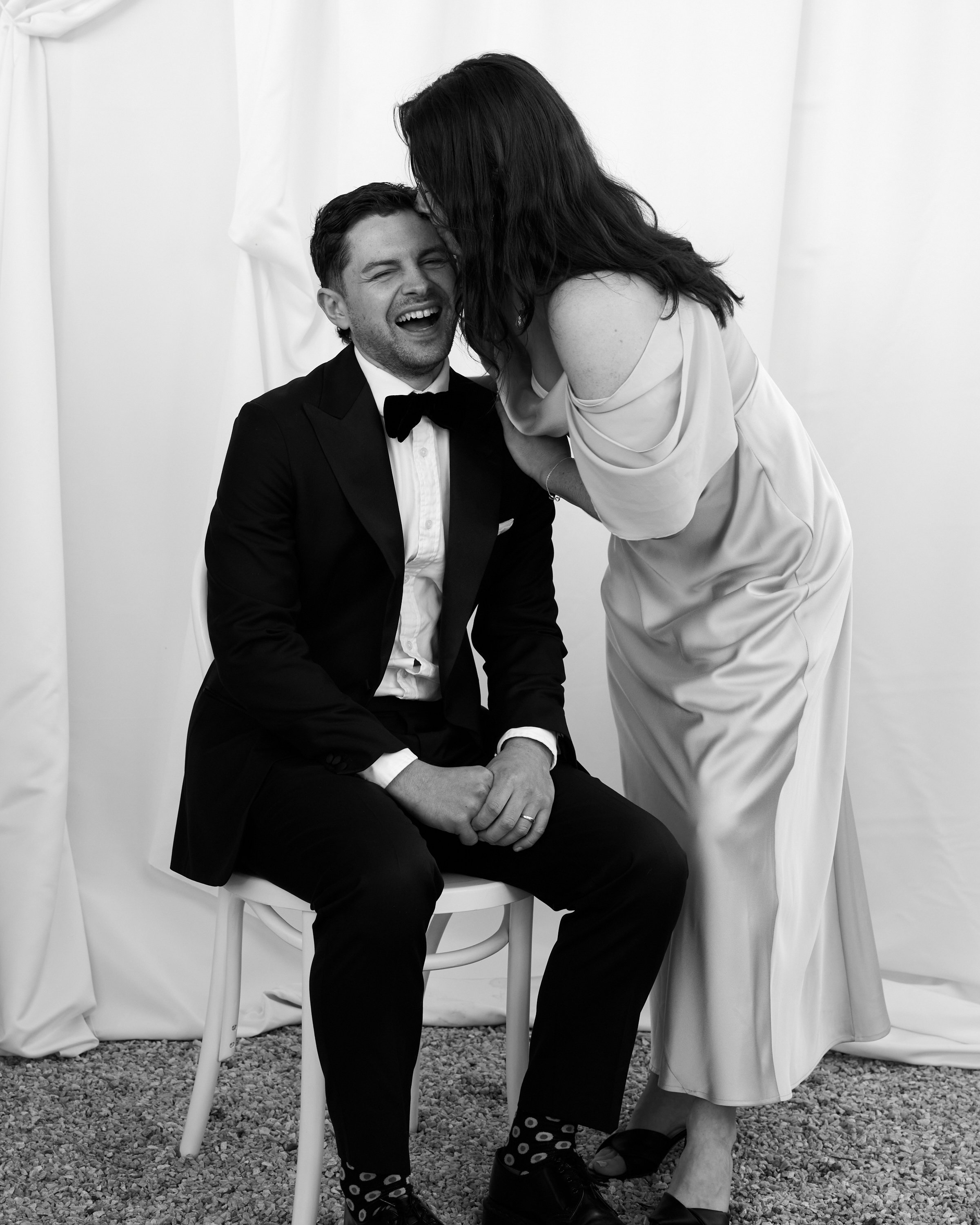 A man in a tuxedo and a woman in an off-shoulder dress sharing a joyful moment; the woman appears to be whispering or kissing the man's head.