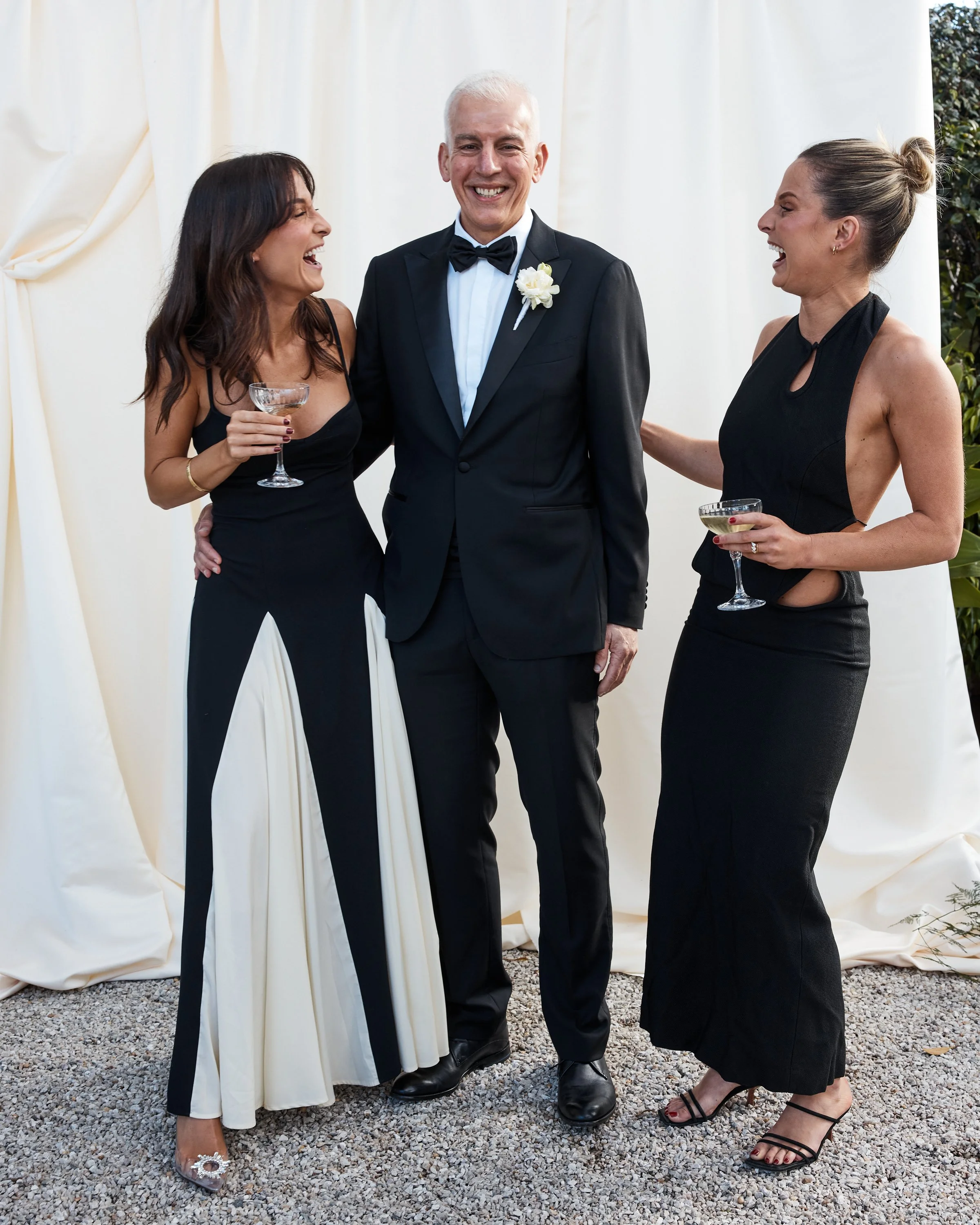 Three people at a wedding celebration, two women in black dresses and a man in a tuxedo, smiling and holding drinks.