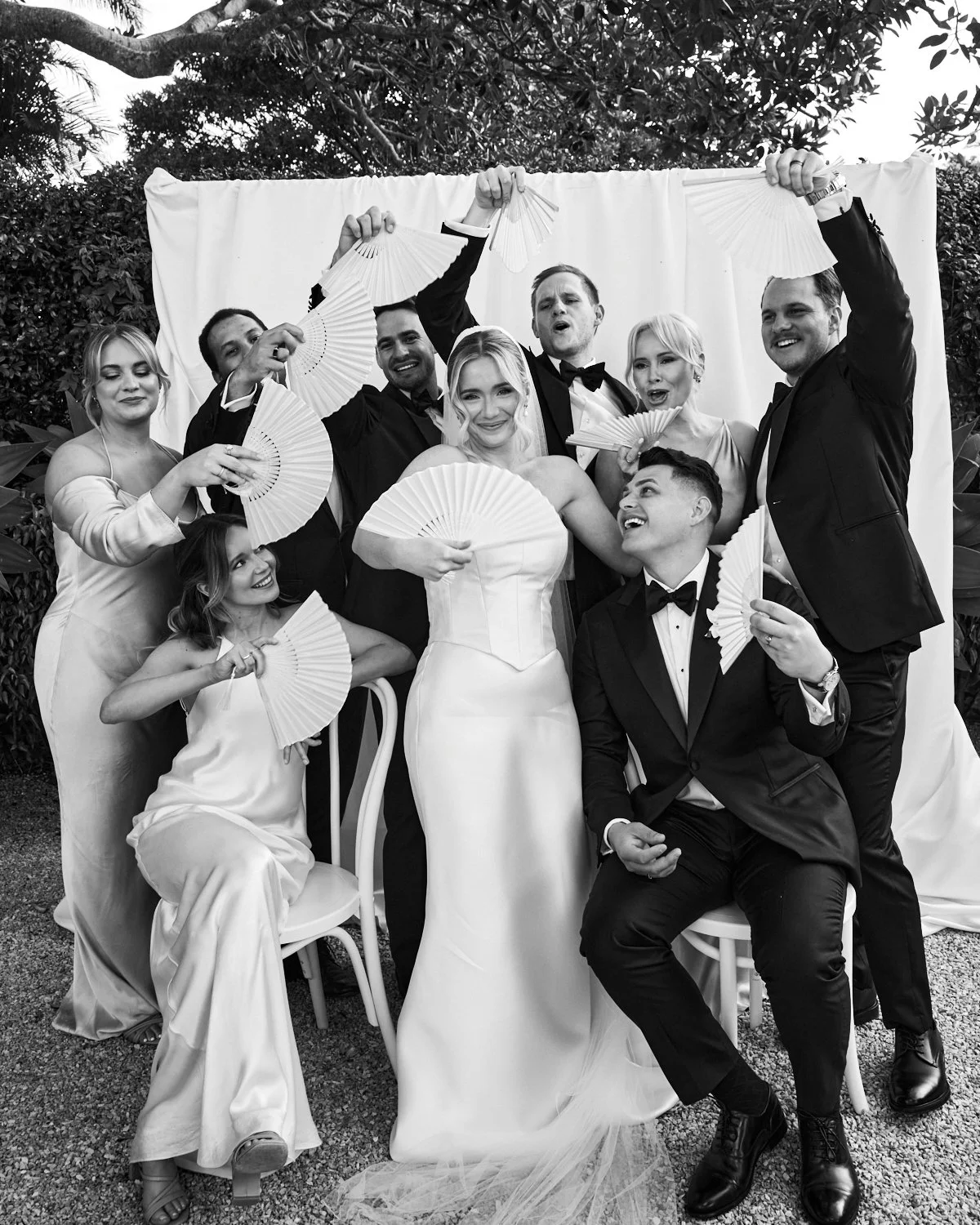 Black and white photo of a wedding party with nine people, including the bride in a strapless wedding dress holding a fan, surrounded by friends and family holding fans, some standing and two seated, outdoors with trees and a white backdrop.