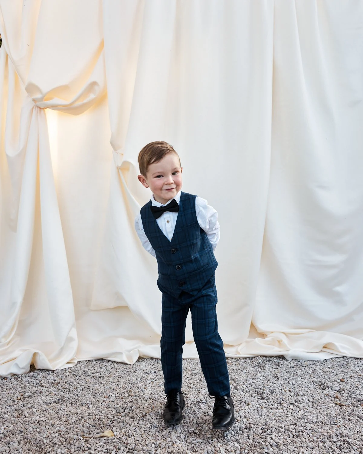 Young boy in a navy blue plaid suit with a white shirt and black bow tie standing on gravel in front of white draped curtains, smiling and looking at the camera.