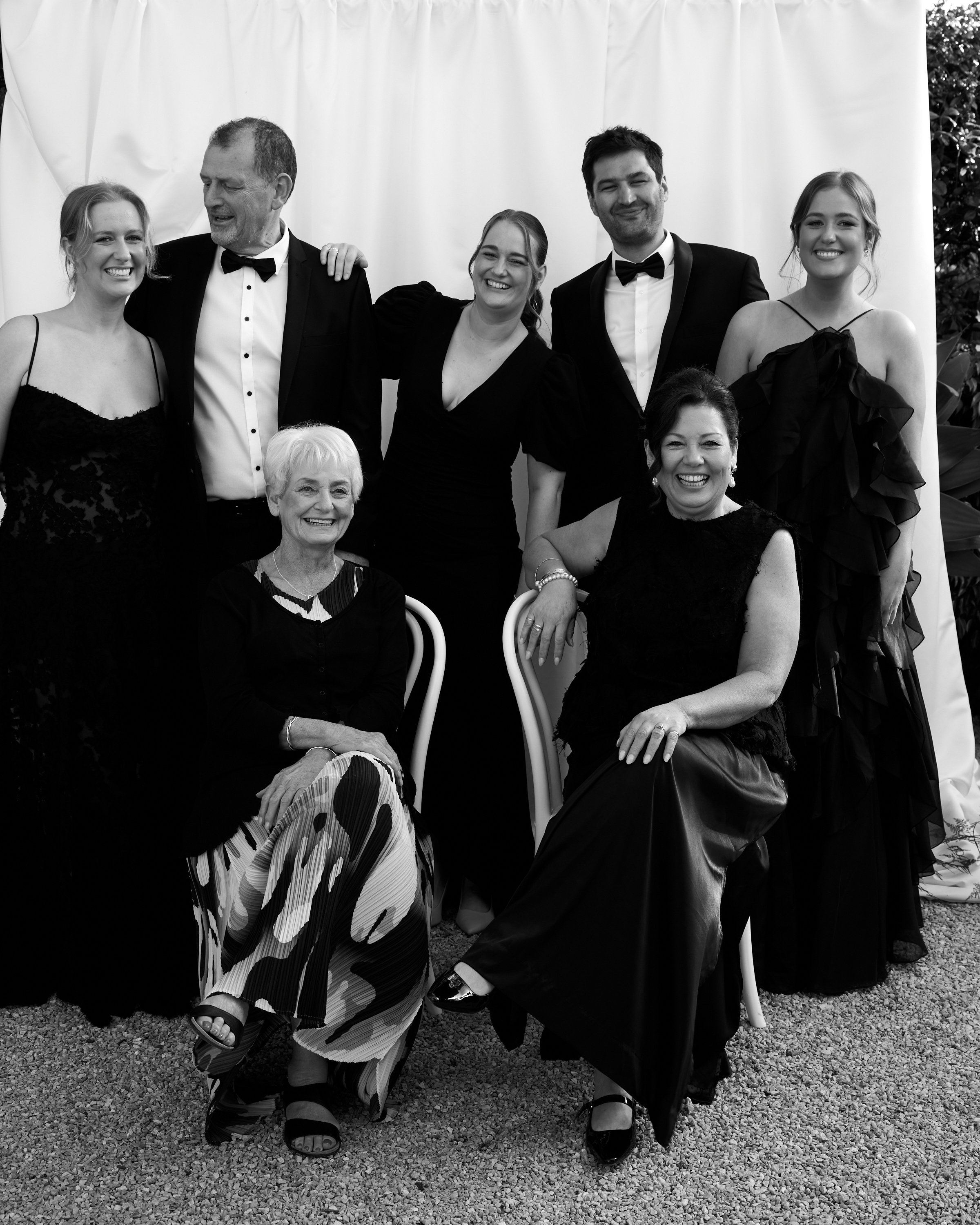 Group of eight people dressed in formal evening attire, posing outdoors in front of a white backdrop, smiling for a photo.