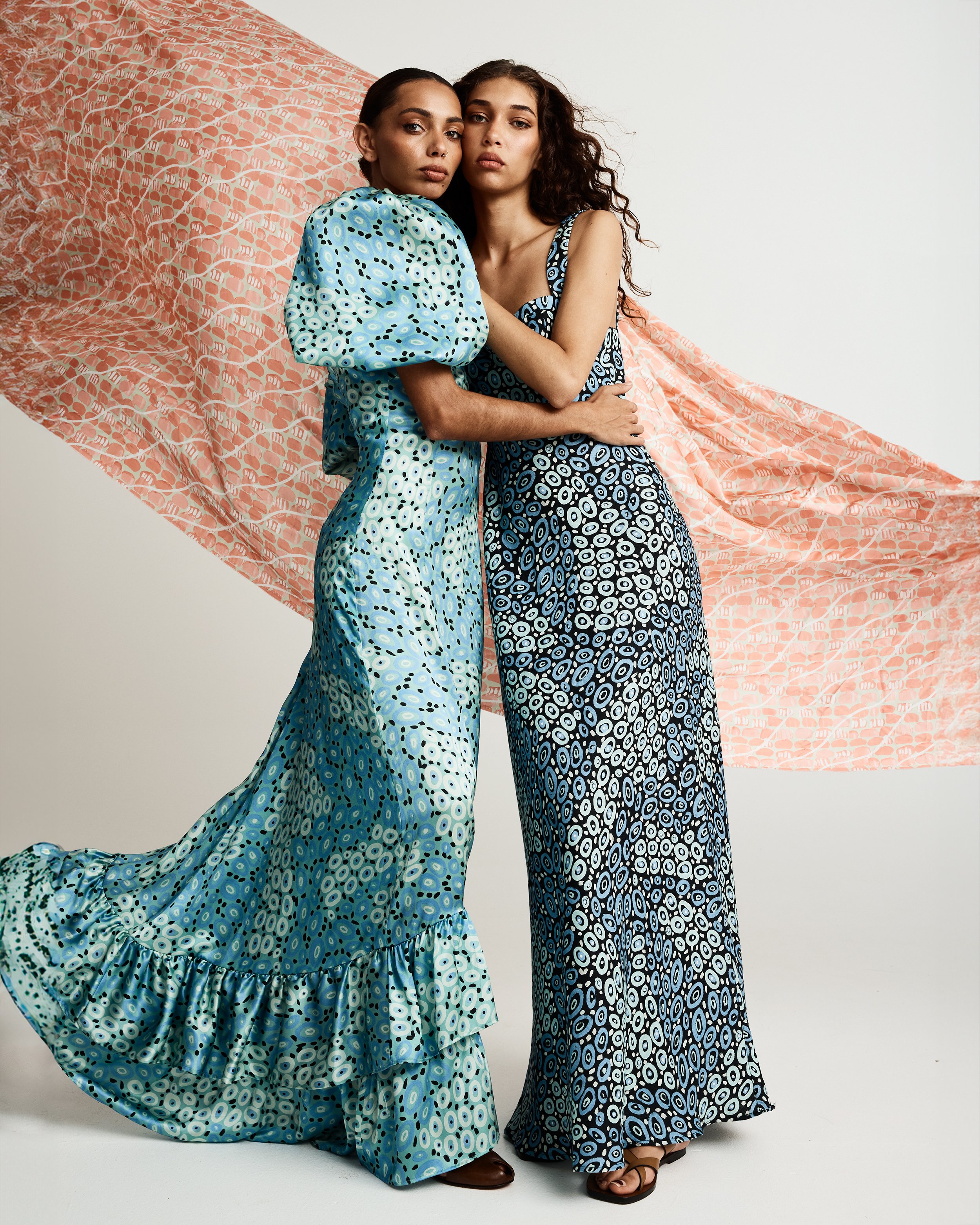 Two women wearing matching patterned dresses standing close together against a backdrop of pink fabric with geometric patterns.