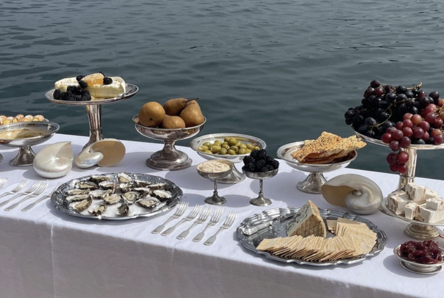 Elegant seafood and fruit spread on a white tablecloth by the water, featuring oysters, grapes, pears, cheeses, crackers, and blackberries.