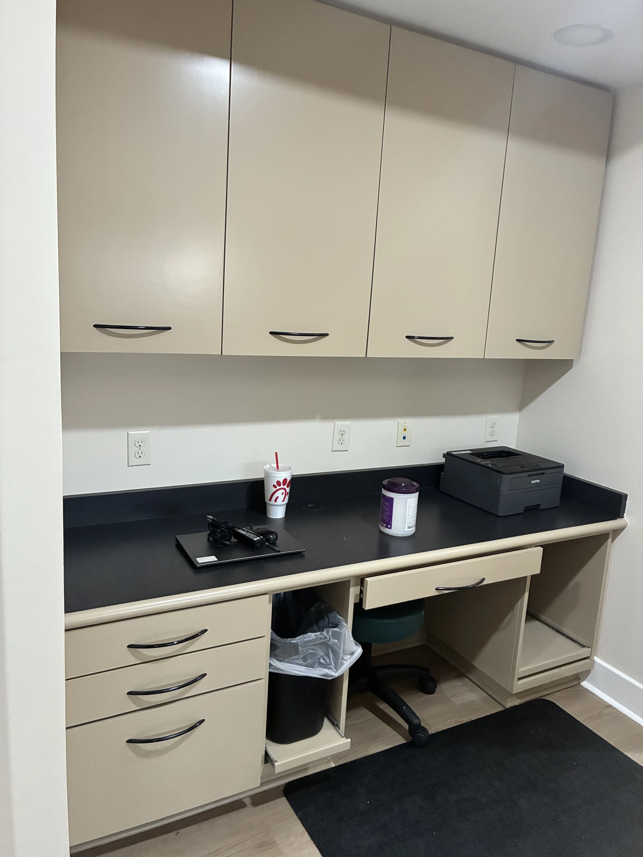 Office kitchen or break area with beige cabinets, a black countertop, a paper cup with a straw, a purple container, a printer, and office supplies on the counter. There's a trash can and a drawer unit under the counter, with electrical outlets on the wall.