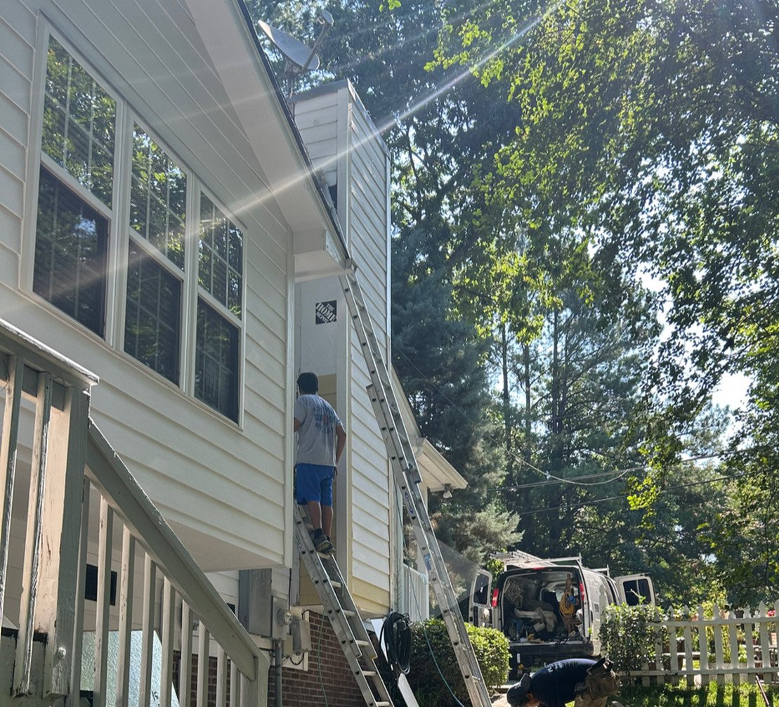Worker on a ladder working on the side of a house, with a van parked nearby and green trees in the background.
