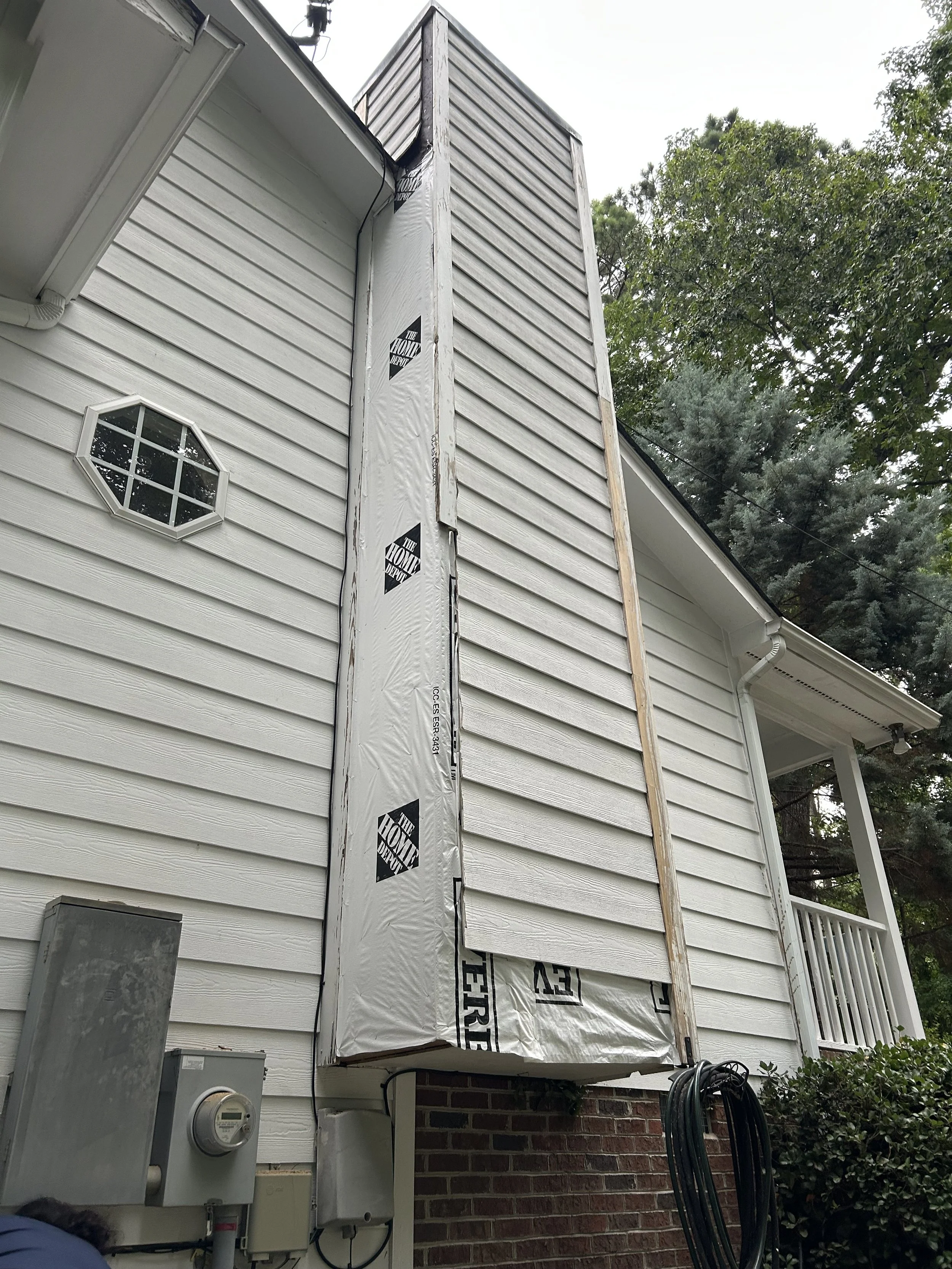 The side of a house with vinyl siding, a small octagonal window, an electrical meter box, and a weatherproofed chimney or vent pipe wrapped in insulation material. There is a wooden post and garden hose on the right side.