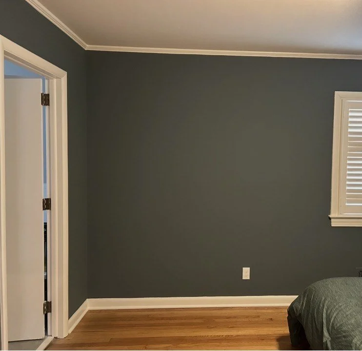 Empty bedroom with dark gray walls, hardwood floor, a small window with white shutters, a white door slightly ajar, and bed in corner.