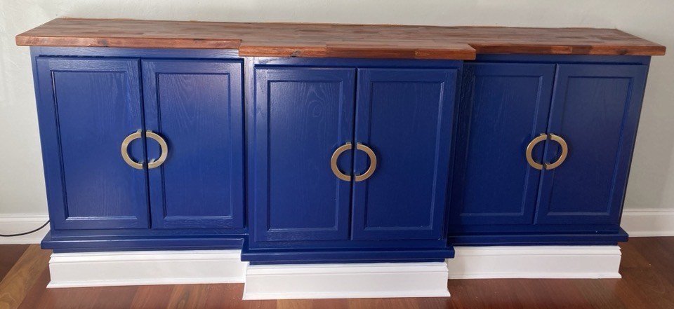 A wooden sideboard painted navy blue with gold ring handles, topped with a natural wood finish surface, placed against a light gray wall with baseboards.
