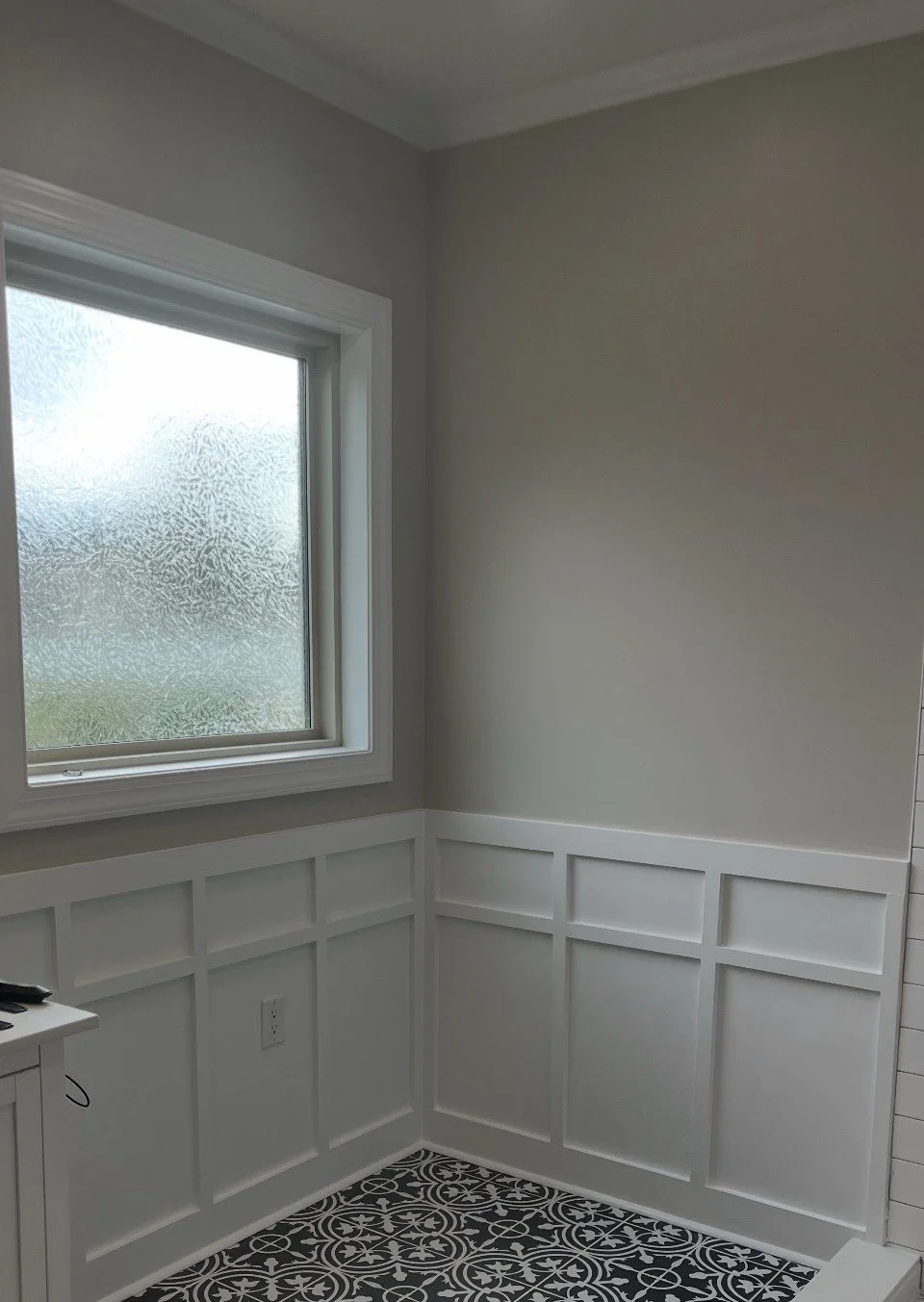 Corner of a room with light gray walls, white wainscoting, a window with frosted glass, and black and white patterned floor tiles.