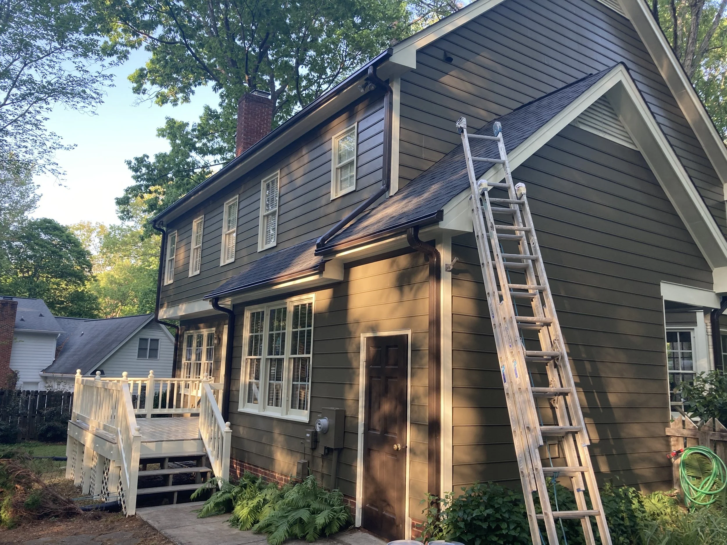 Two-story house with dark green siding, white trim, and multiple large windows, situated in a leafy neighborhood with trees and other houses visible in the background. A metal ladder leans against the house near the roof, and there's a small wooden deck with stairs in the backyard.