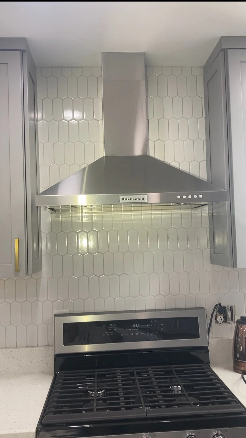 Kitchen with a stainless steel range hood above a stove, gray cabinets, and a white backsplash with hexagonal tiles.