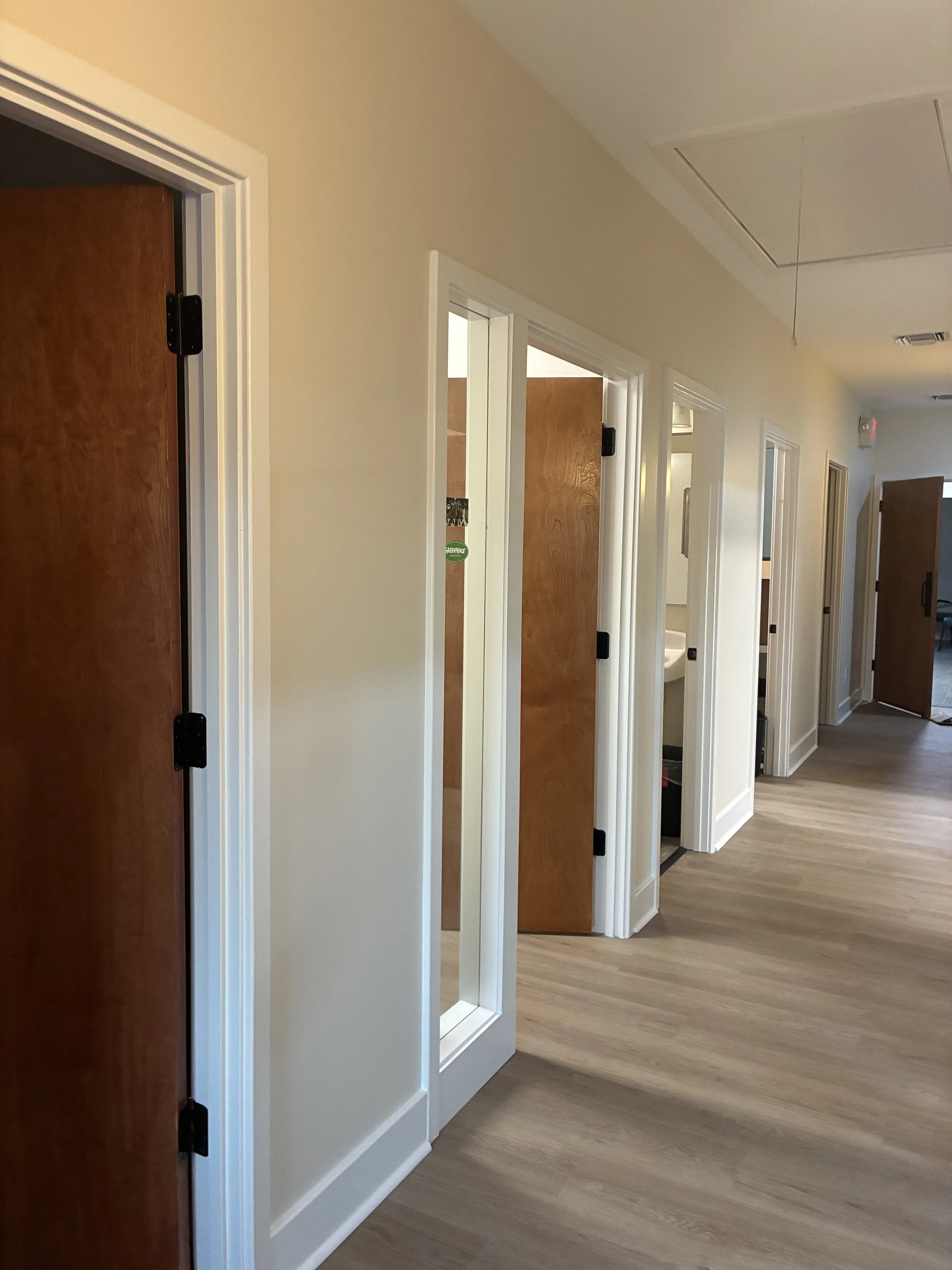 A hallway with multiple doors on the right side, some of which are open, revealing interior rooms. The hallway has light-colored walls, white door frames, and wood-like flooring. There is a ceiling hatch and a hanging string or cord in the ceiling.