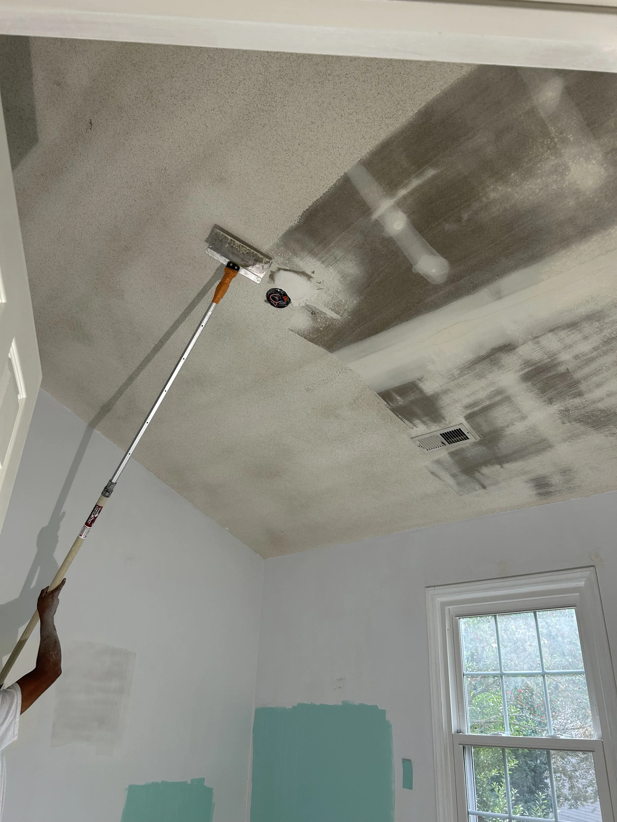 Person painting a room ceiling with a paint roller, with partially painted walls and a window showing trees outside.