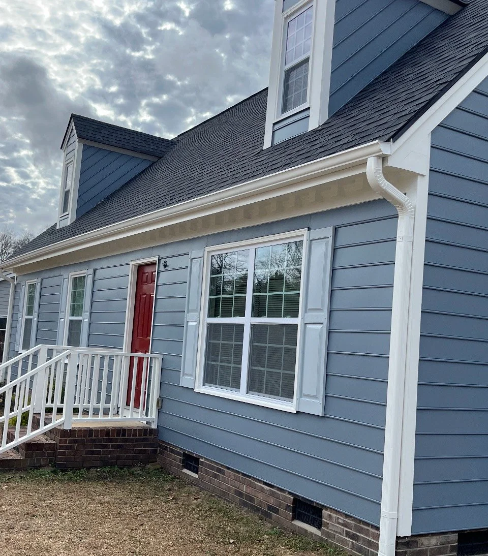 Blue house with white trim, brick foundation, white railing stairs, red front door, three windows with white shutters, and a divided light design, under a partly cloudy sky.