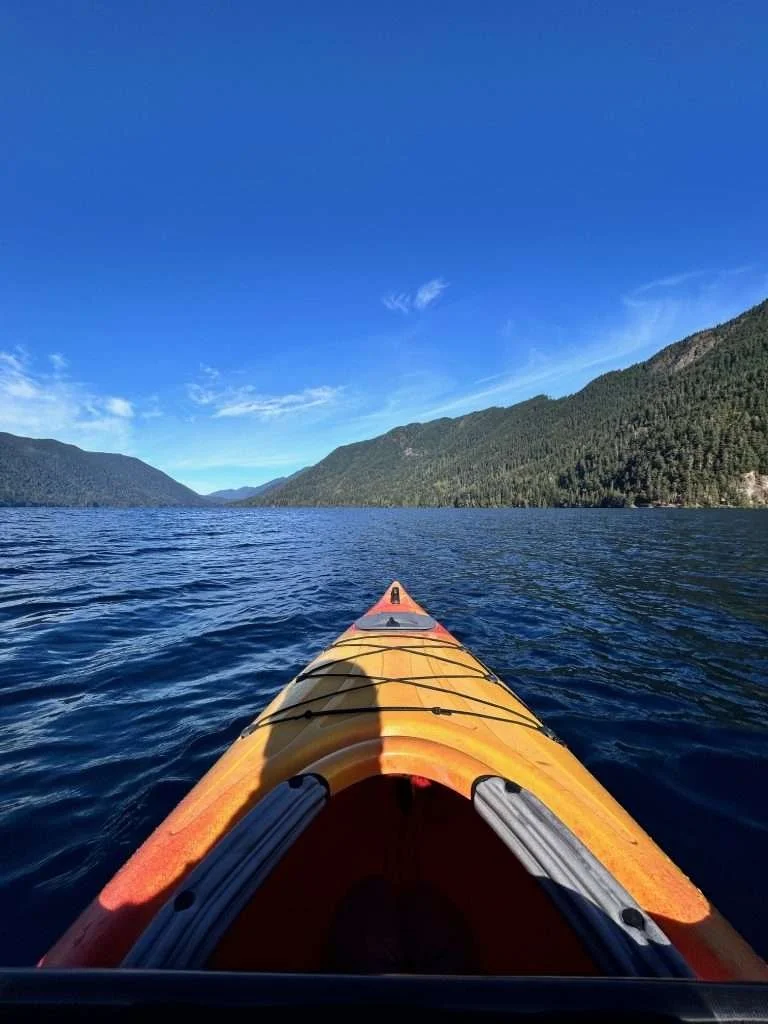 Orange kayak on a calm lake with green forested mountains in the background under a clear blue sky.