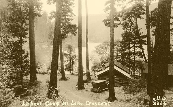 Black and white photo of a wooded area at Lake Crescent with a campsite, including tents and vintage cars, and a small cabin among tall trees.