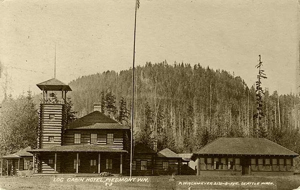 Historical black and white photo of the Log Cabin Hotel in Piedmont, Washington, with a wooden tower, a main building, and the forested hills in the background.