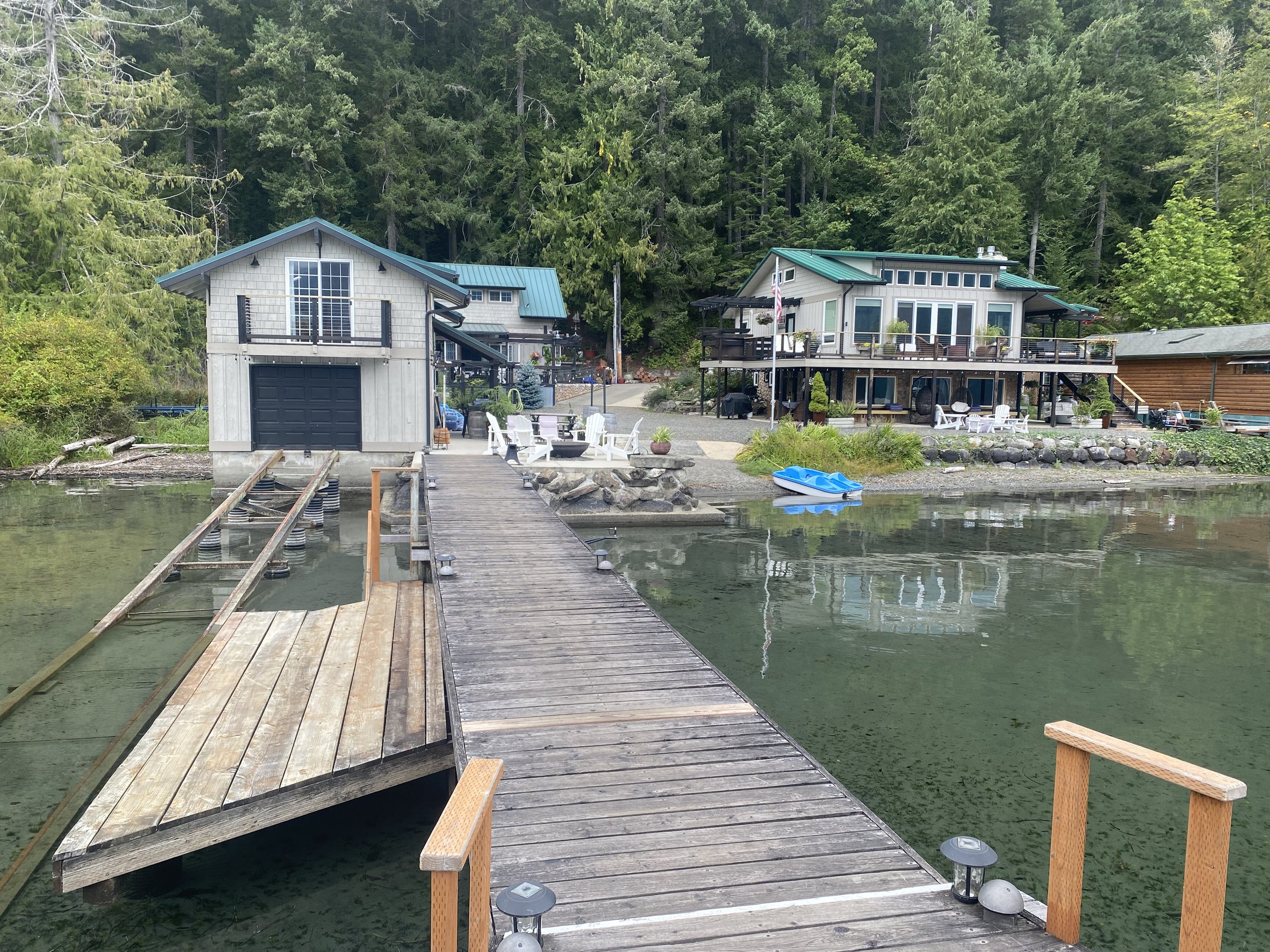 A wooden dock extending into a calm body of water, with a small blue boat tied to the right side, leading to two houses on a tree-lined shore. One house is on stilts with large windows and a deck, and the other has a garage and outdoor seating area.