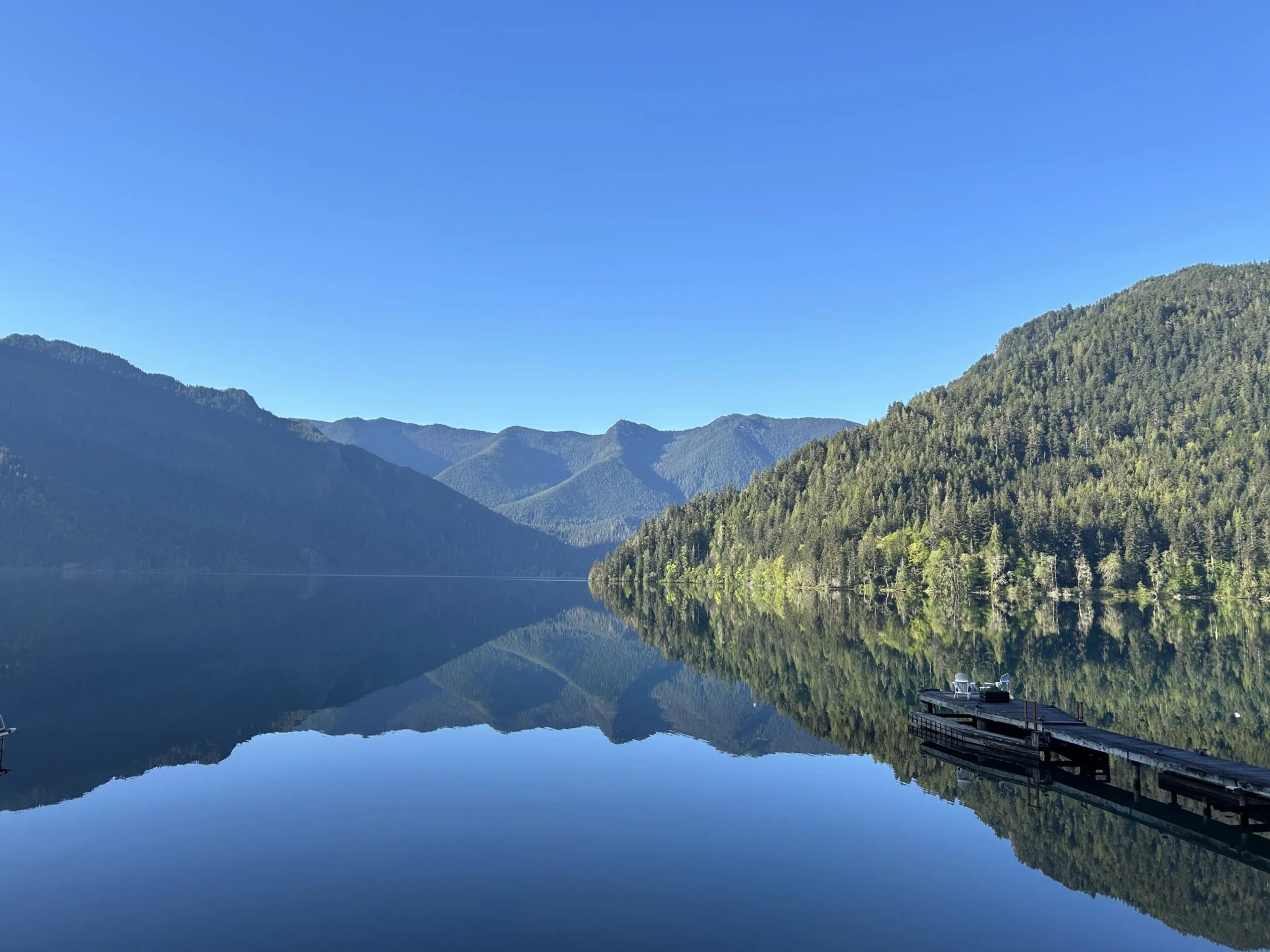 Calm lake reflecting surrounding green forested mountains under a clear blue sky.