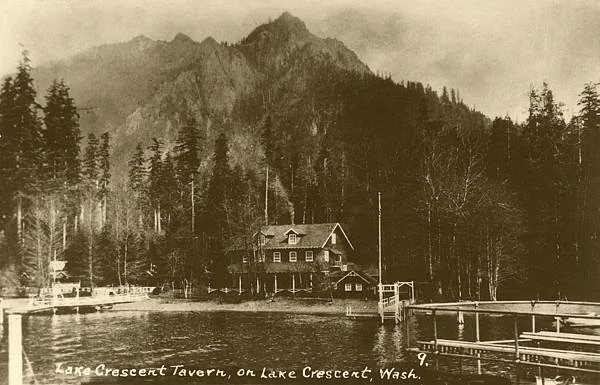 A sepia-toned photograph of Lake Crescent Tavern on Lake Crescent, Washington, with a mountain range and dense trees in the background, and a wooden dock extending into the water.
