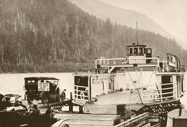 A vintage ferry boat docked at a pier with several cars and people nearby, set against a backdrop of forested hills and mountains.