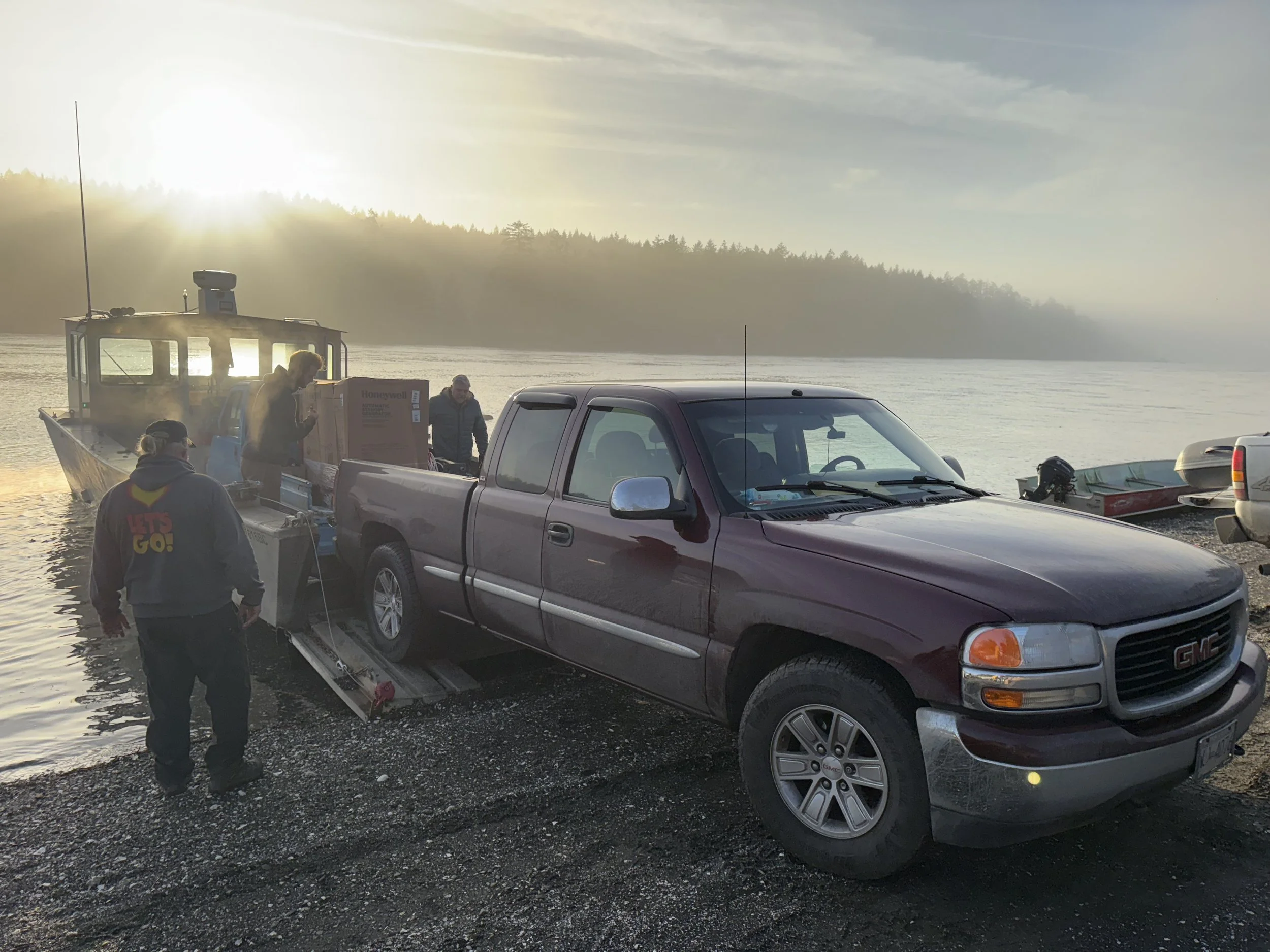 A maroon GMC pickup truck on a gravel shore with a boat ramp, being loaded onto a boat trailer by a group of people, with a boat in the water and boats on the shore, during sunrise or sunset.