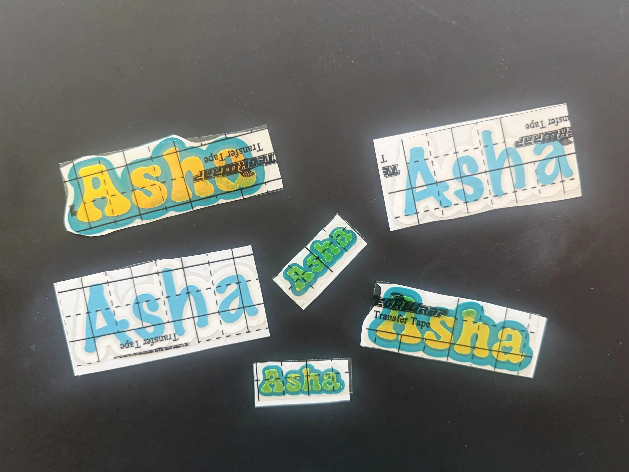 Various colorful name tags and stickers with the name 'Asha' written on them, in different fonts and styles, placed on a dark surface.