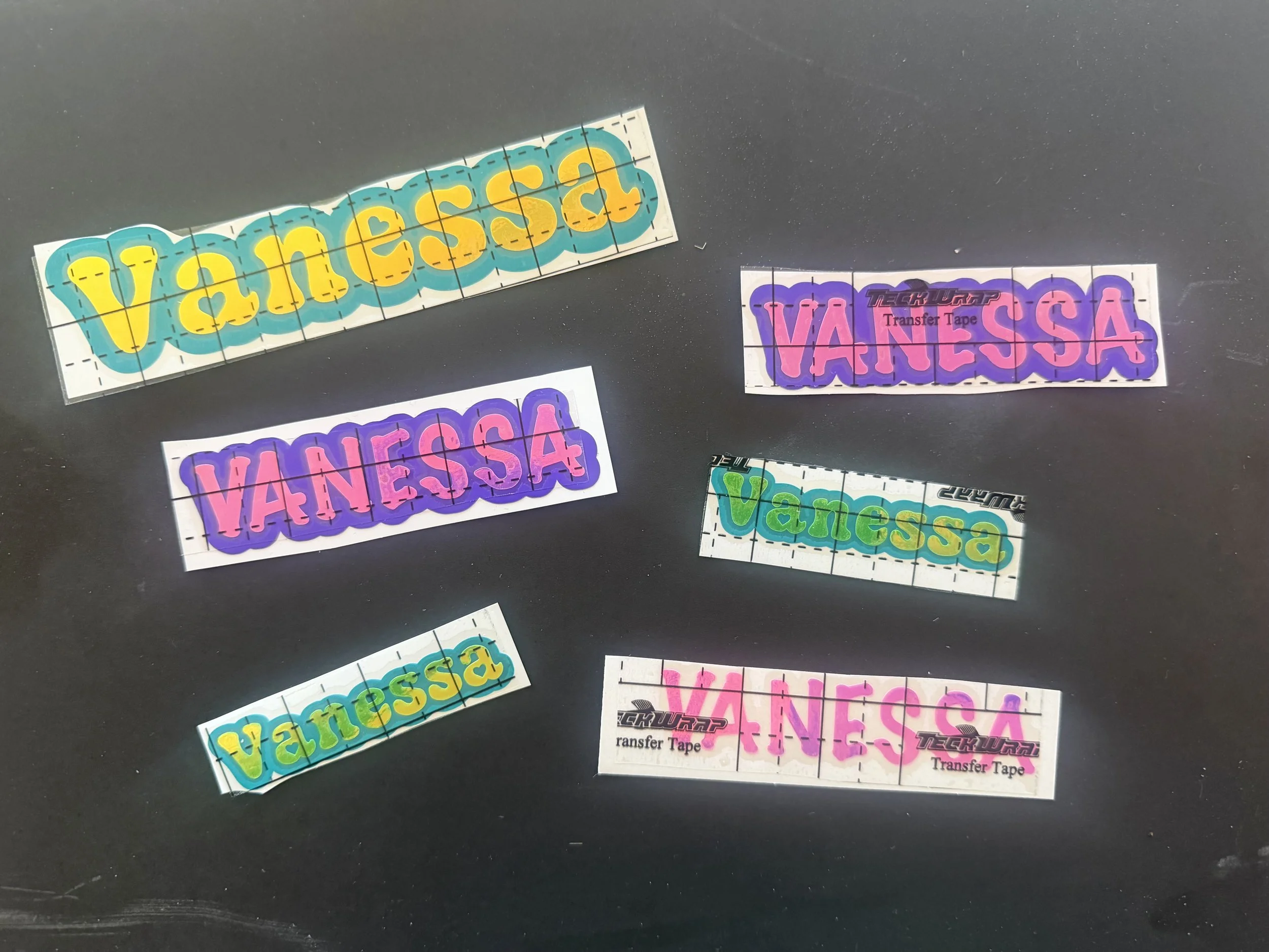 Multiple colorful stickers with the word Vanessa in different fonts and colors, some with a background grid and others with included branding, spread on a black surface.