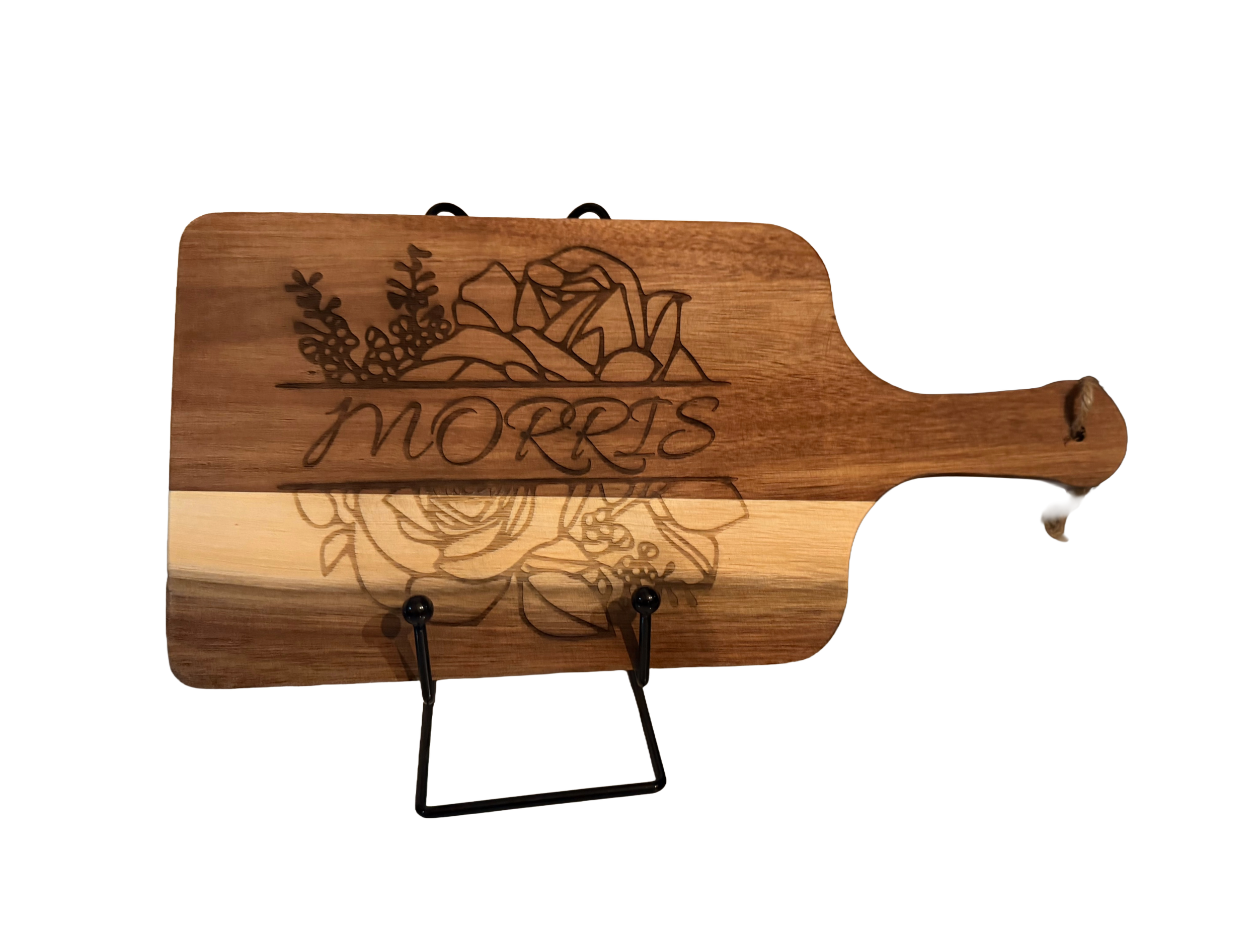 Wooden cutting board with floral engraving and the word 'MORRIS' written across it, displayed on a black metal stand with a black background.