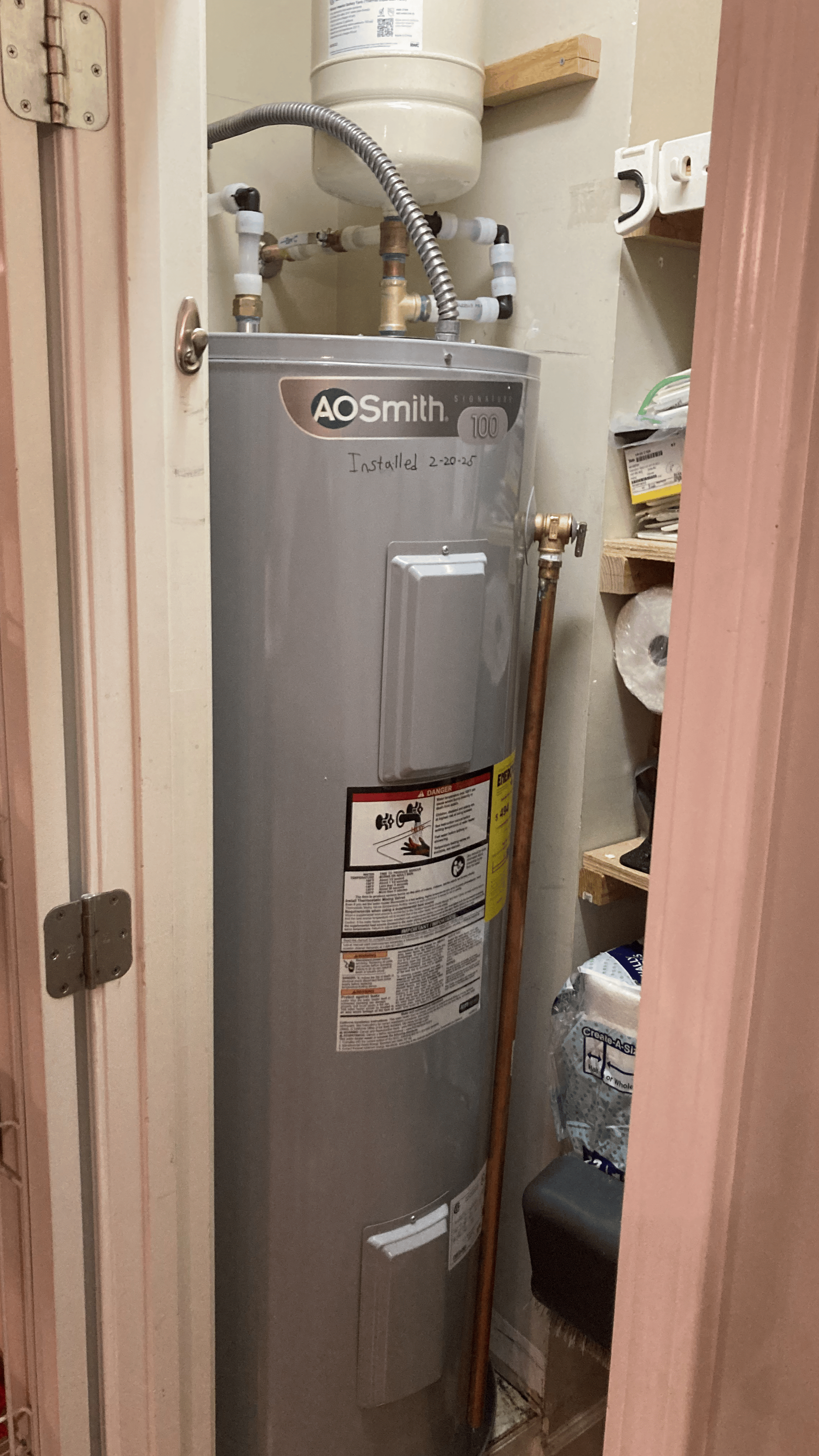 A water heater in a small closet with pipes connected on top and a broom leaning against it.