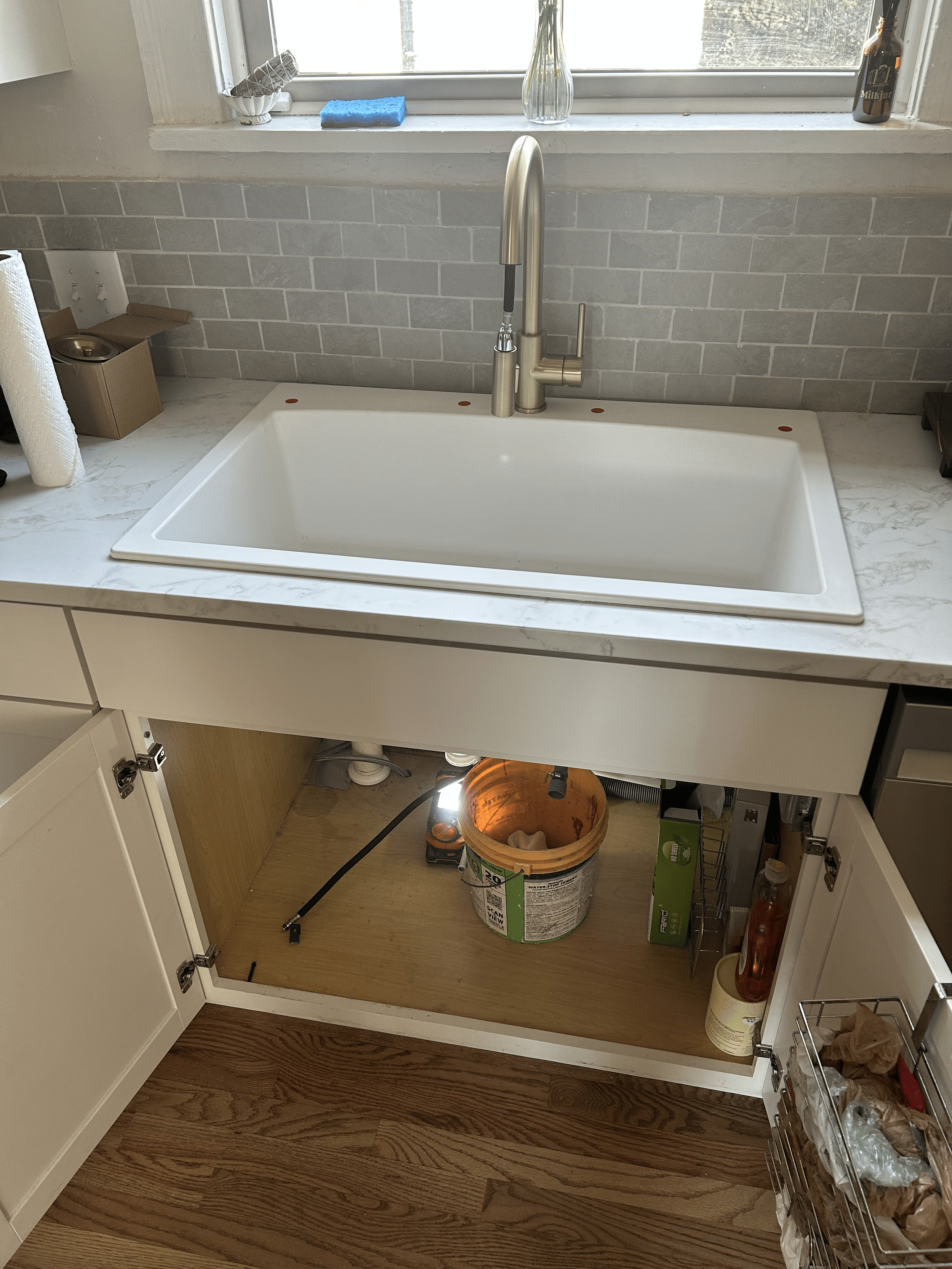 Kitchen sink with a modern faucet, window above, and an open cabinet below containing a bucket, a power tool, and some other items. Countertop has a roll of paper towels, a small box, and a soap bottle. The backsplash is made of light gray subway tiles.