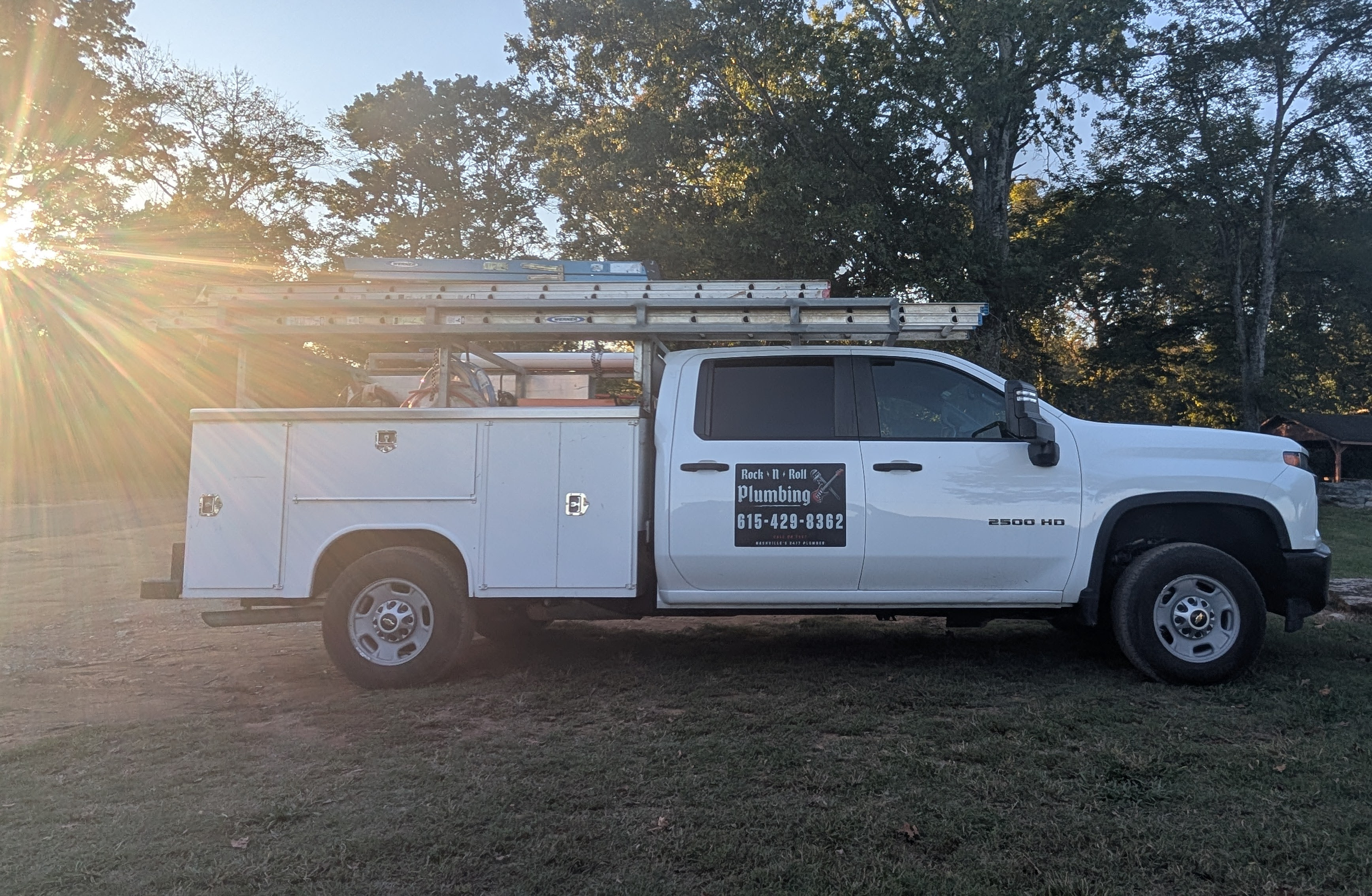 White plumbing service truck with ladders on top, parked on grass with trees in the background during sunset.