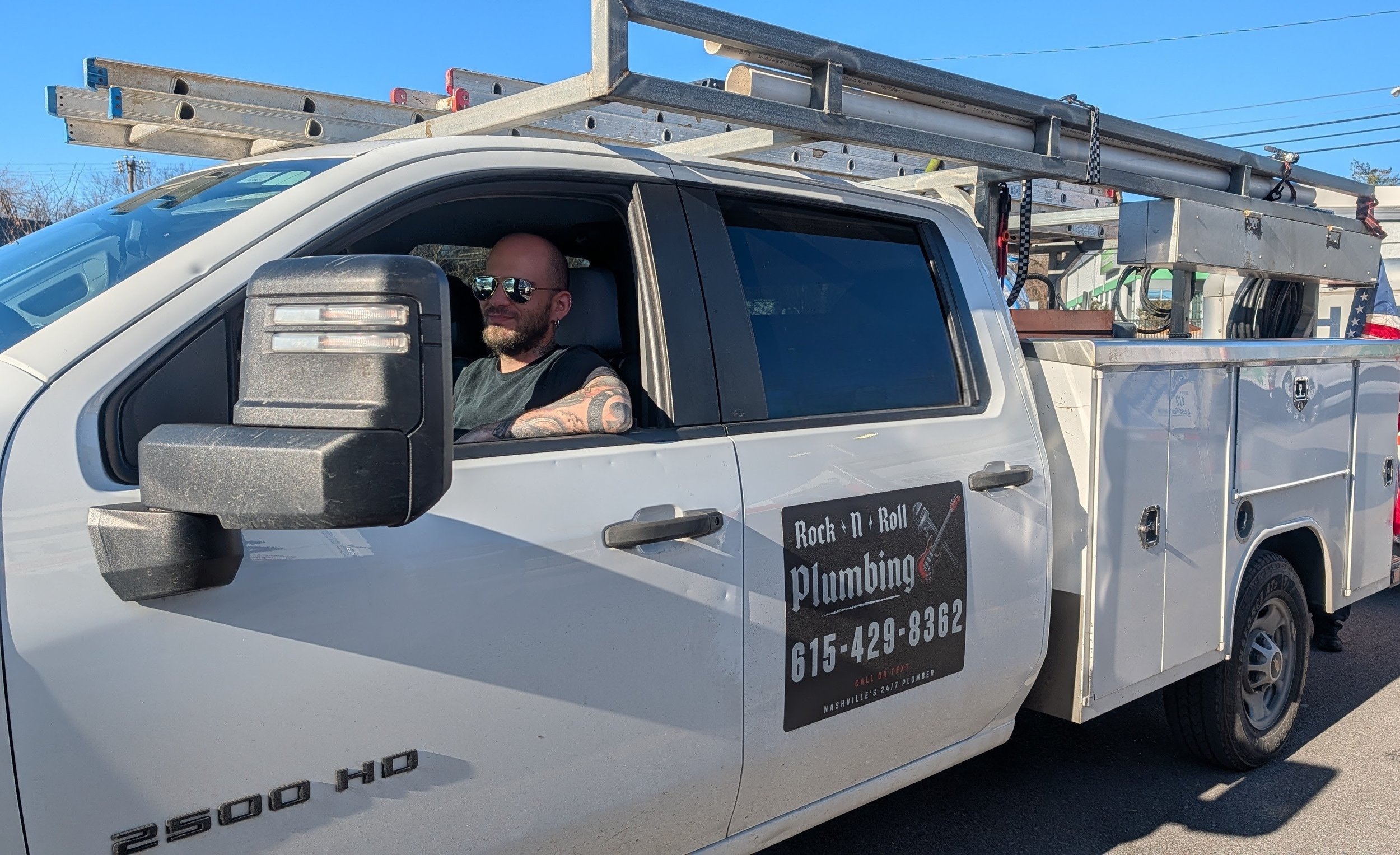 A white service pickup truck with a man wearing sunglasses sitting in the driver’s seat. The truck has ladder and equipment storage in the back, with ladders mounted on top. The door has a sign for a plumbing company that reads "Rock N Roll Plumbing" with a phone number, 615-429-8362.