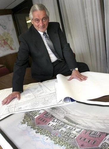 An elderly man in a suit and striped tie is leaning on a table with architectural plans and a colored drawing of a building project.