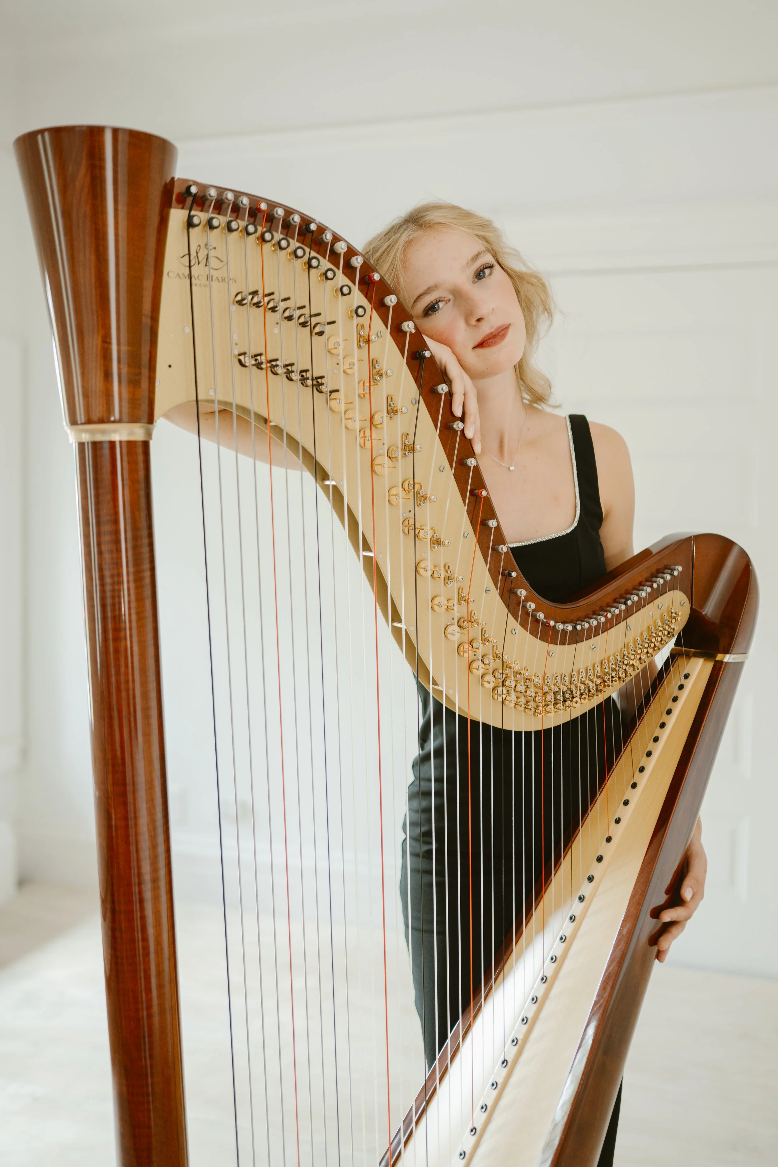 Young woman with blonde hair posing with a large wooden harp in a bright, minimal room.