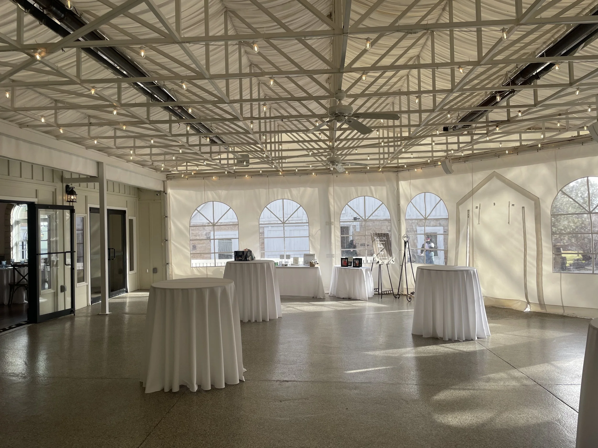 An empty event space with a high ceiling adorned with string lights, round tables draped with white tablecloths, large arched windows allowing natural light, and some display stands near the back wall.