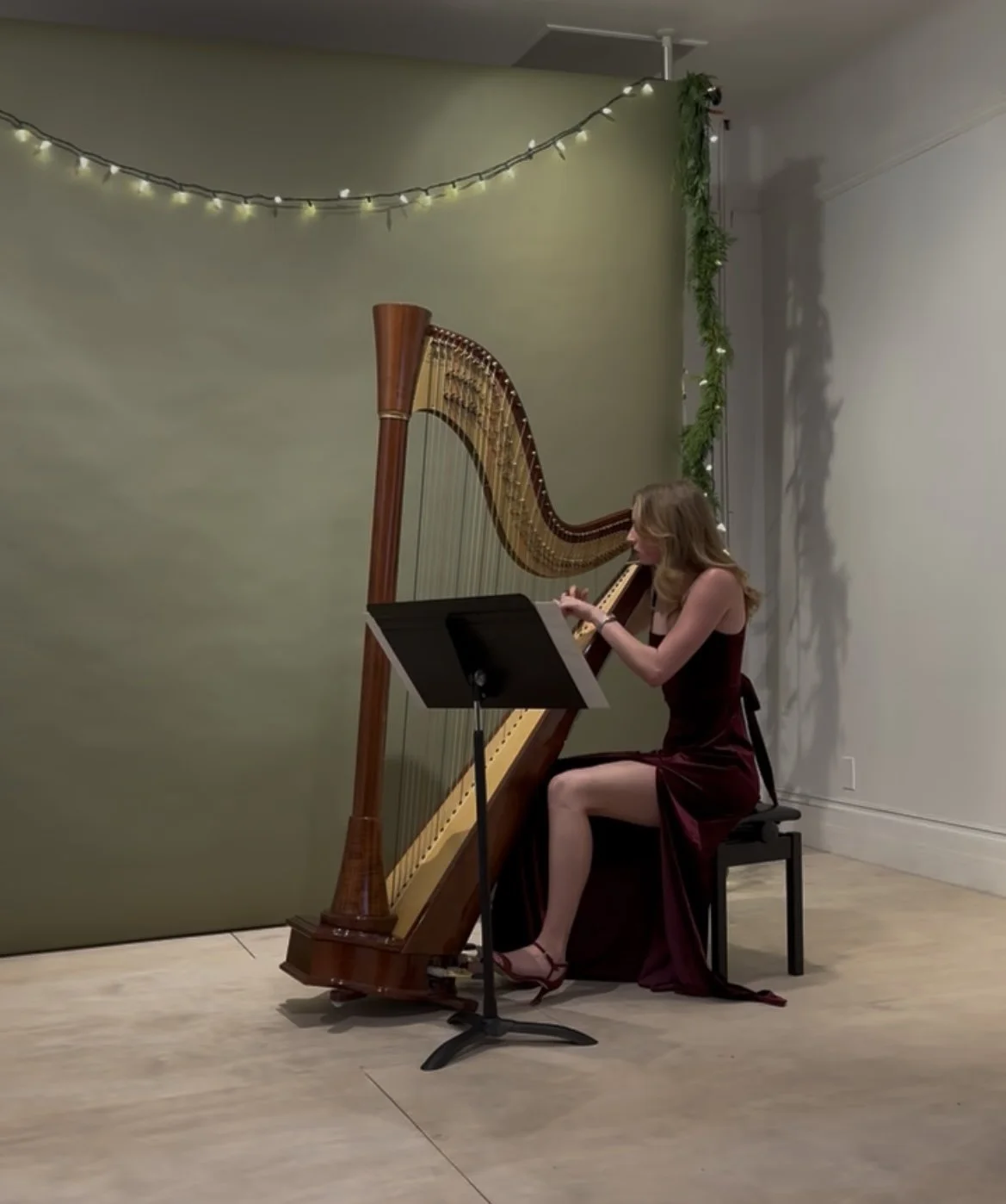 A woman in a burgundy dress playing a harp on stage with a music stand, string lights, and greenery decoration.
