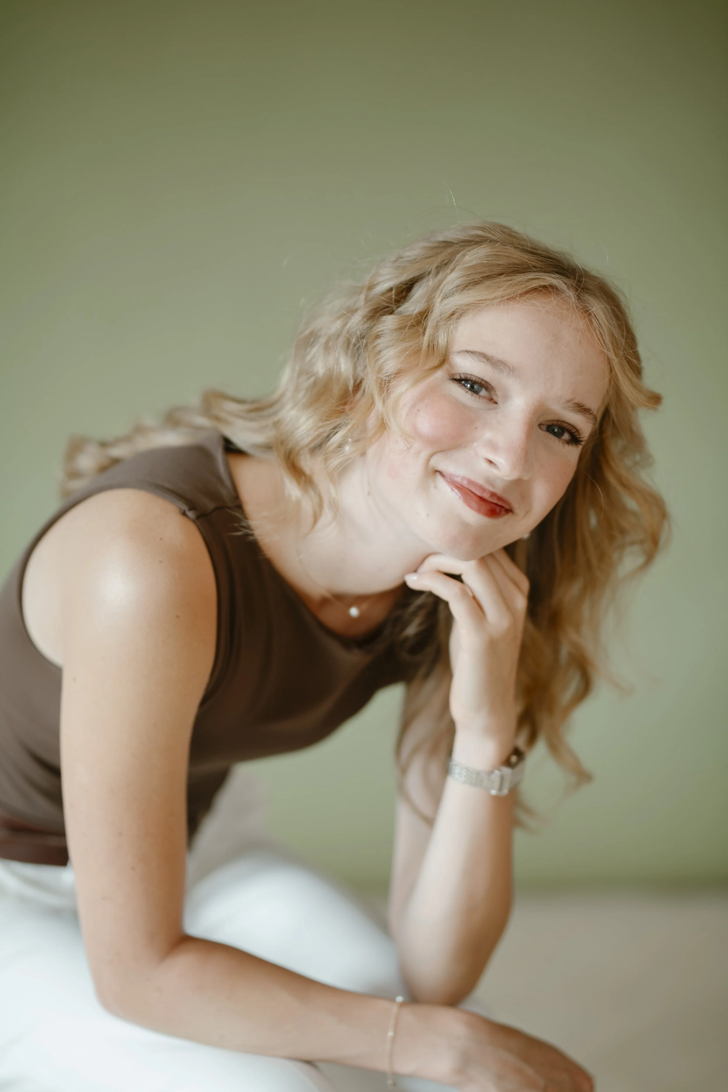 A young woman with blonde hair and a brown sleeveless top, smiling and resting her chin on her hand against a light green background.