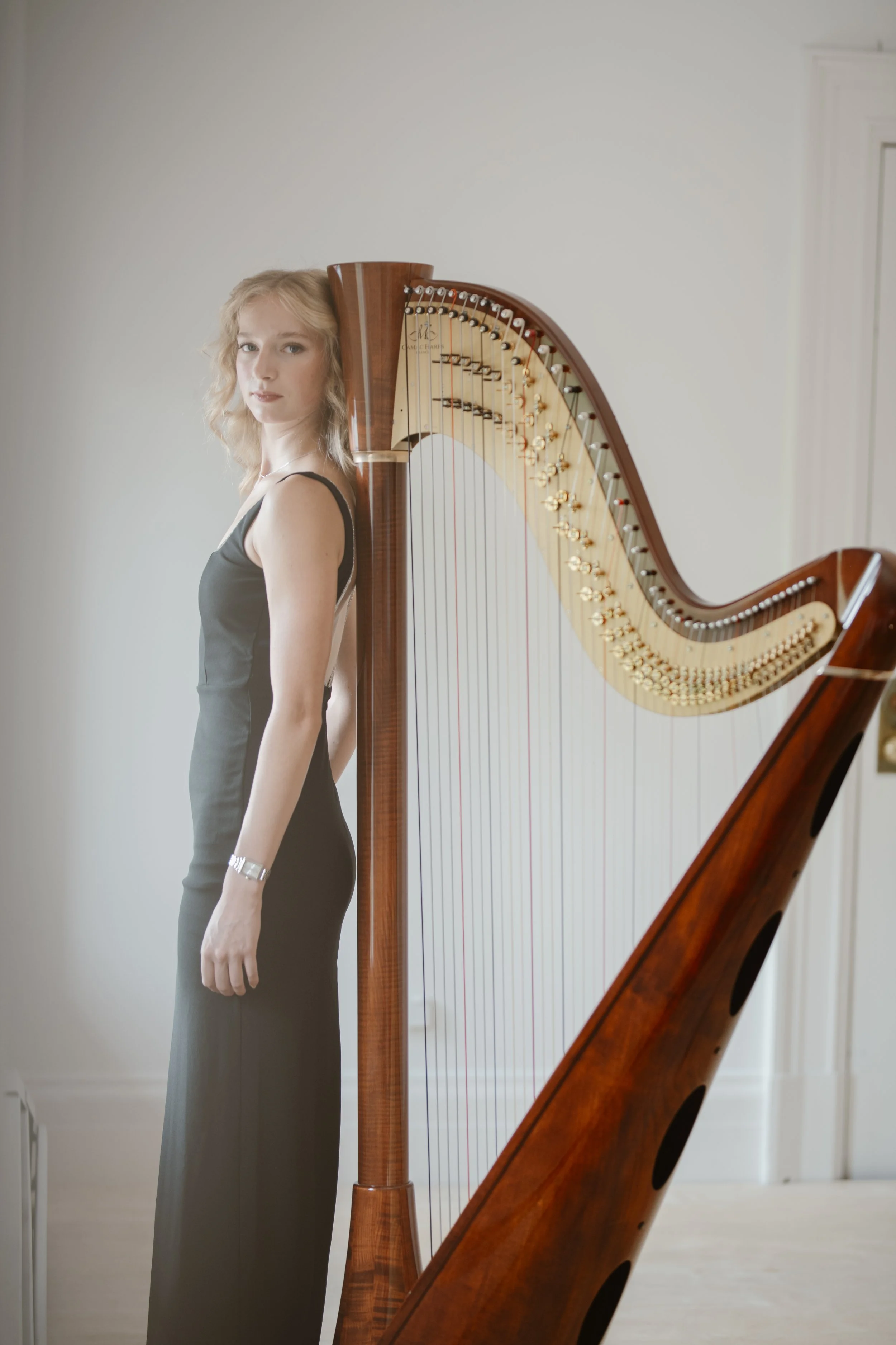 Young woman in a black dress standing next to a wooden harp in a room.