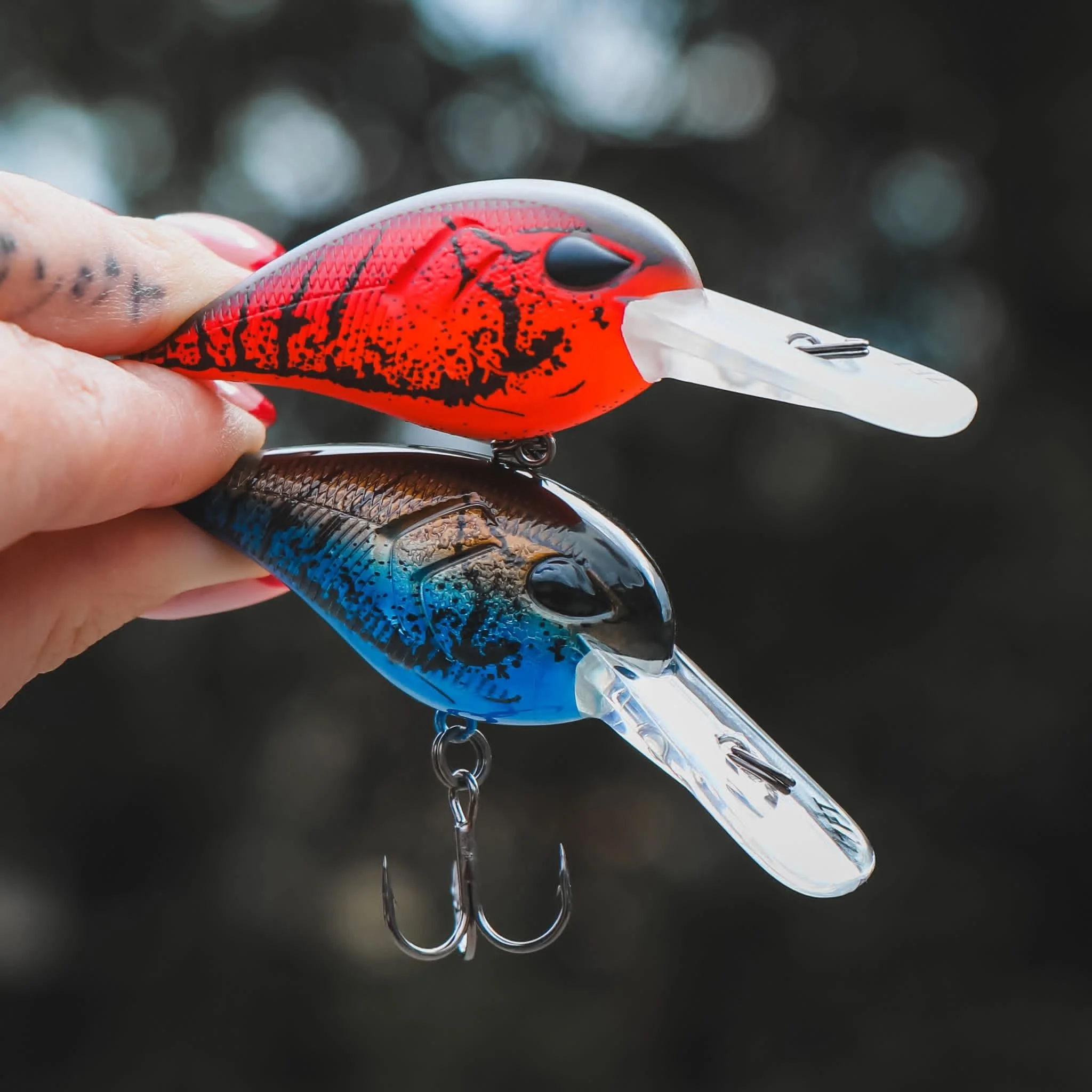 Two fishing lures, one red and black, and one black and blue, held in a person's hand with a dark, blurred background.